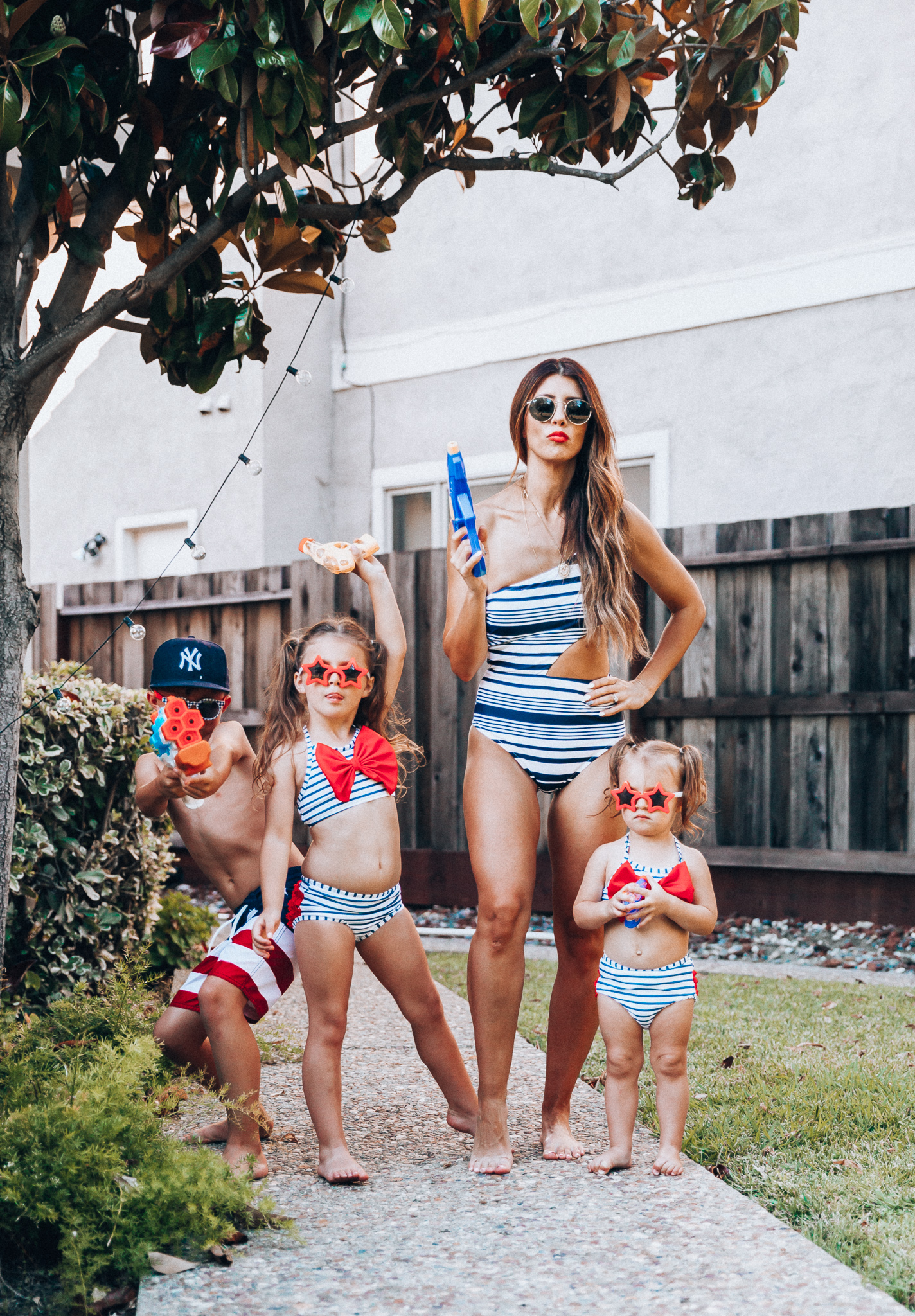 Water Gun Fights + Cute Summer Swimsuits for the Family by popular San Francisco fashion blog, The Girl in the Yellow Dress: image of a woman, two young girls and young boy standing outside holding water guns and wearing red, white and blue RuffleButts Little Girls Bikini 2-Piece Swimsuit with Bow and Ruffles, star and grinderPUNCH Kids American USA Flag Sunglasses, ON-FIELD COLLECTION Kids/Youth Yankees 59Fifty Fitted Navy Hat Cap, Old Navy American flag Patterned Board Shorts for Boys, navy Aerie Cutout one shoulder one piece swimsuit, and Nordstrom Ray-Ban Icons 53mm Retro Sunglasses.