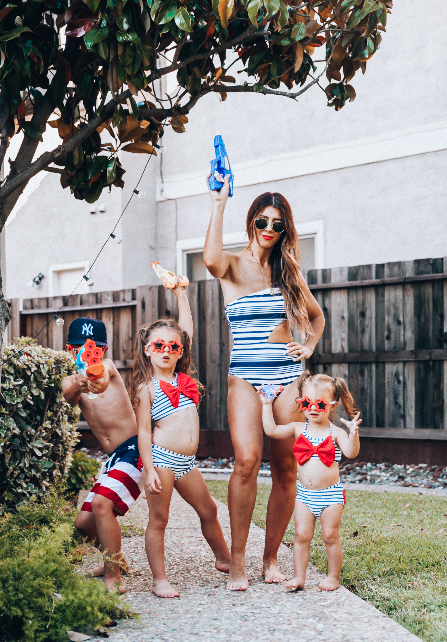 Water Gun Fights + Cute Summer Swimsuits for the Family by popular San Francisco fashion blog, The Girl in the Yellow Dress: image of a woman, two young girls and young boy standing outside holding water guns and wearing red, white and blue RuffleButts Little Girls Bikini 2-Piece Swimsuit with Bow and Ruffles, star and grinderPUNCH Kids American USA Flag Sunglasses, ON-FIELD COLLECTION Kids/Youth Yankees 59Fifty Fitted Navy Hat Cap, Old Navy American flag Patterned Board Shorts for Boys, navy Aerie Cutout one shoulder one piece swimsuit, and Nordstrom Ray-Ban Icons 53mm Retro Sunglasses.