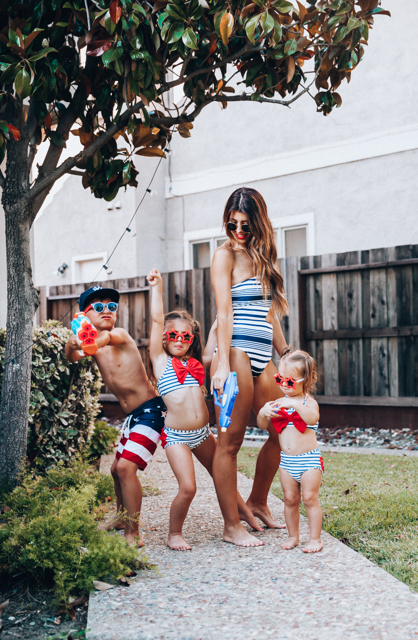 Water Gun Fights + Cute Summer Swimsuits for the Family by popular San Francisco fashion blog, The Girl in the Yellow Dress: image of a woman, two young girls and young boy standing outside holding water guns and wearing red, white and blue RuffleButts Little Girls Bikini 2-Piece Swimsuit with Bow and Ruffles, star and grinderPUNCH Kids American USA Flag Sunglasses, ON-FIELD COLLECTION Kids/Youth Yankees 59Fifty Fitted Navy Hat Cap, Old Navy American flag Patterned Board Shorts for Boys, navy Aerie Cutout one shoulder one piece swimsuit, and Nordstrom Ray-Ban Icons 53mm Retro Sunglasses.