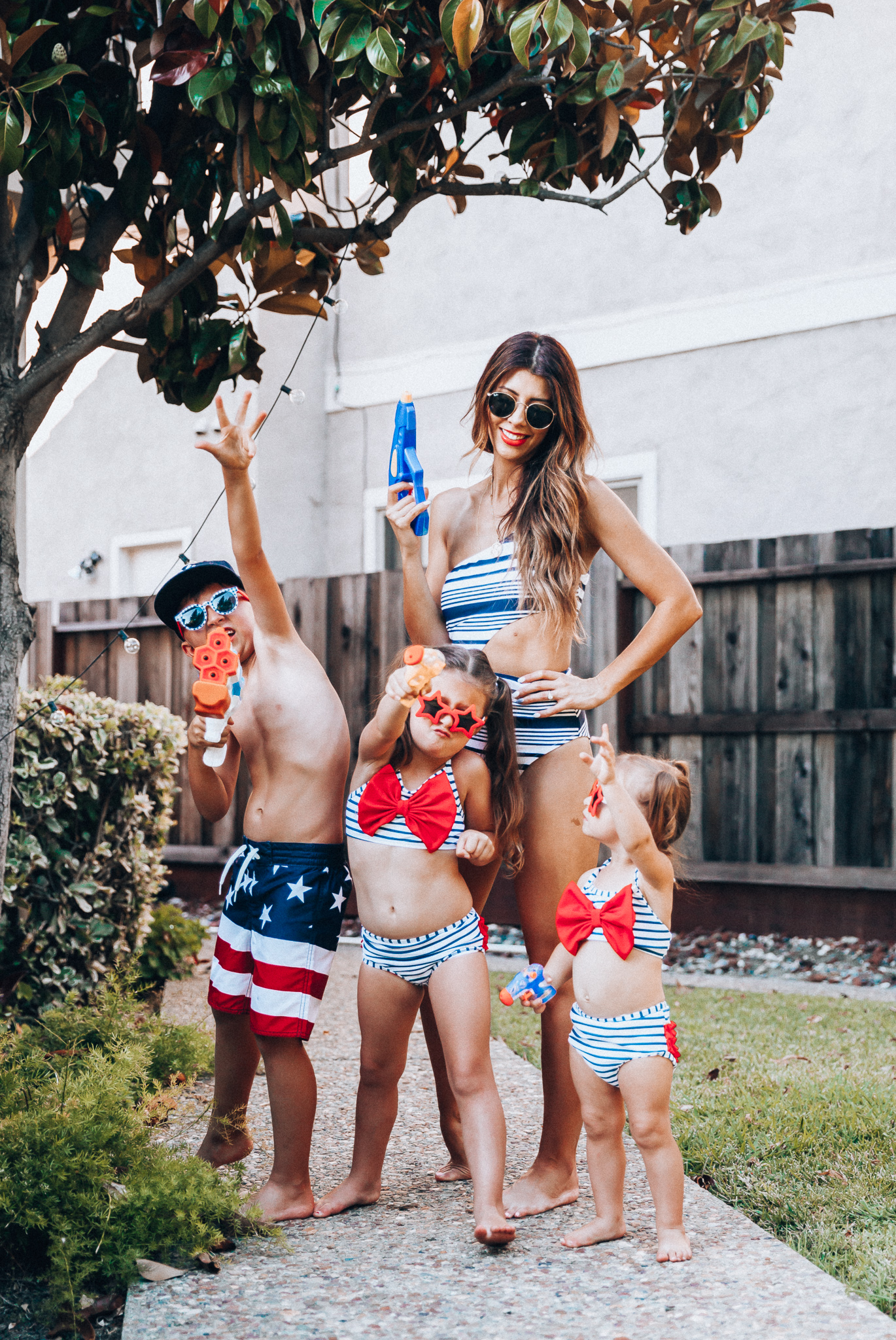 Water Gun Fights + Cute Summer Swimsuits for the Family by popular San Francisco fashion blog, The Girl in the Yellow Dress: image of a woman, two young girls and young boy standing outside holding water guns and wearing red, white and blue RuffleButts Little Girls Bikini 2-Piece Swimsuit with Bow and Ruffles, star and grinderPUNCH Kids American USA Flag Sunglasses, ON-FIELD COLLECTION Kids/Youth Yankees 59Fifty Fitted Navy Hat Cap, Old Navy American flag Patterned Board Shorts for Boys, navy Aerie Cutout one shoulder one piece swimsuit, and Nordstrom Ray-Ban Icons 53mm Retro Sunglasses.