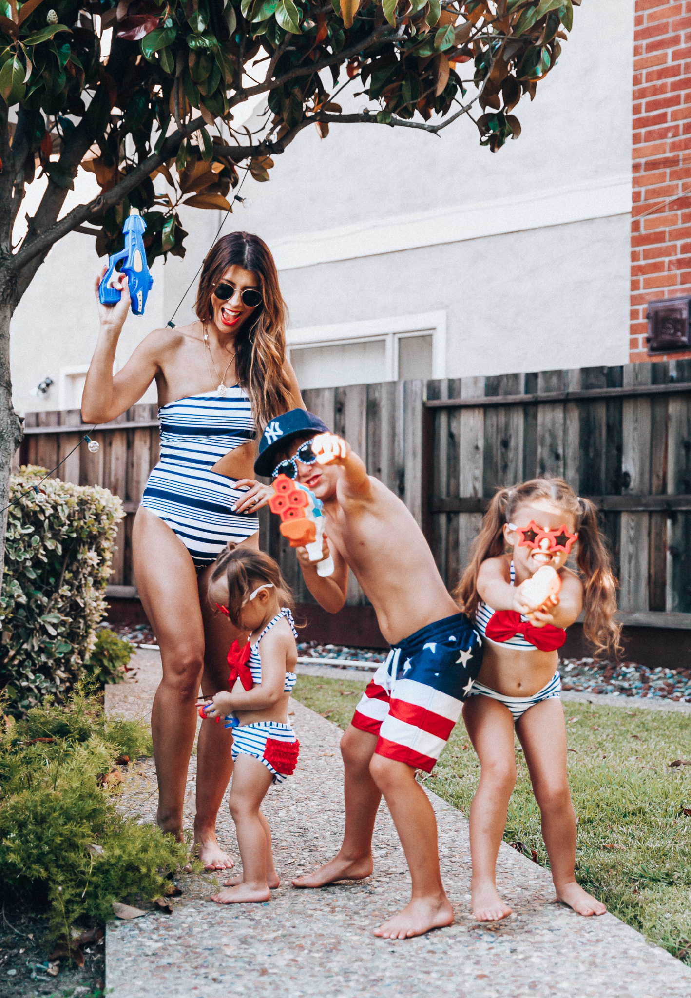 Water Gun Fights + Cute Summer Swimsuits for the Family by popular San Francisco fashion blog, The Girl in the Yellow Dress: image of a woman, two young girls and young boy standing outside holding water guns and wearing red, white and blue RuffleButts Little Girls Bikini 2-Piece Swimsuit with Bow and Ruffles, star and grinderPUNCH Kids American USA Flag Sunglasses, ON-FIELD COLLECTION Kids/Youth Yankees 59Fifty Fitted Navy Hat Cap, Old Navy American flag Patterned Board Shorts for Boys, navy Aerie Cutout one shoulder one piece swimsuit, and Nordstrom Ray-Ban Icons 53mm Retro Sunglasses.