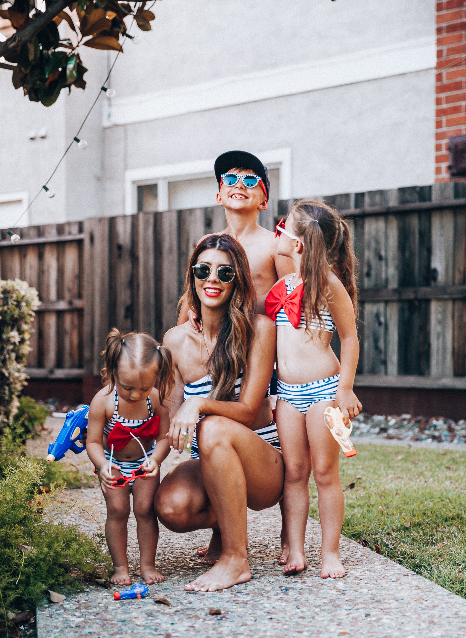 Water Gun Fights + Cute Summer Swimsuits for the Family by popular San Francisco fashion blog, The Girl in the Yellow Dress: image of a woman, two young girls and young boy standing outside holding water guns and wearing red, white and blue RuffleButts Little Girls Bikini 2-Piece Swimsuit with Bow and Ruffles, star and grinderPUNCH Kids American USA Flag Sunglasses, ON-FIELD COLLECTION Kids/Youth Yankees 59Fifty Fitted Navy Hat Cap, Old Navy American flag Patterned Board Shorts for Boys, navy Aerie Cutout one shoulder one piece swimsuit, and Nordstrom Ray-Ban Icons 53mm Retro Sunglasses.