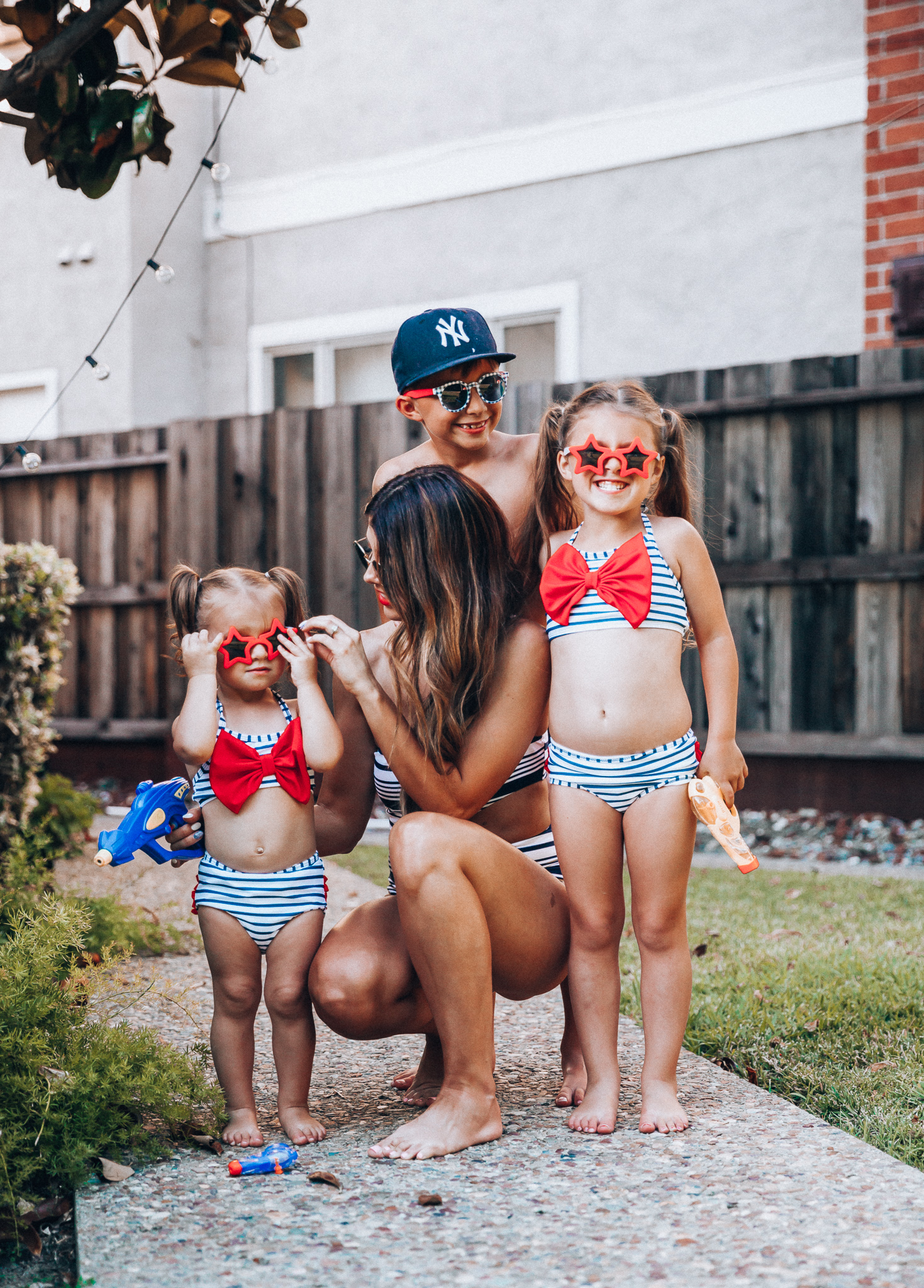 Water Gun Fights + Cute Summer Swimsuits for the Family by popular San Francisco fashion blog, The Girl in the Yellow Dress: image of a woman, two young girls and young boy standing outside holding water guns and wearing red, white and blue RuffleButts Little Girls Bikini 2-Piece Swimsuit with Bow and Ruffles, star and grinderPUNCH Kids American USA Flag Sunglasses, ON-FIELD COLLECTION Kids/Youth Yankees 59Fifty Fitted Navy Hat Cap, Old Navy American flag Patterned Board Shorts for Boys, navy Aerie Cutout one shoulder one piece swimsuit, and Nordstrom Ray-Ban Icons 53mm Retro Sunglasses.