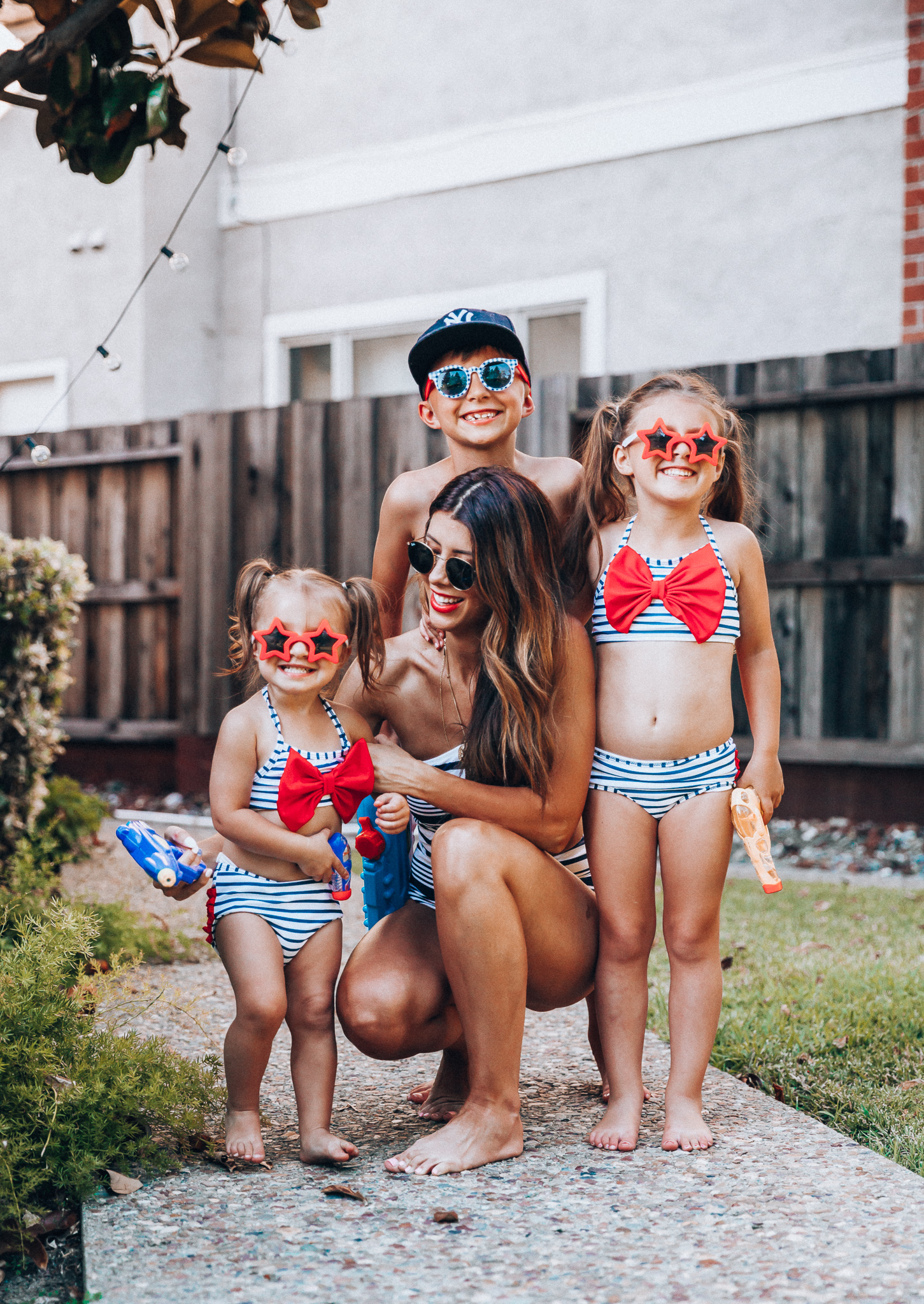 Water Gun Fights + Cute Summer Swimsuits for the Family by popular San Francisco fashion blog, The Girl in the Yellow Dress: image of a woman, two young girls and young boy standing outside holding water guns and wearing red, white and blue RuffleButts Little Girls Bikini 2-Piece Swimsuit with Bow and Ruffles, star and grinderPUNCH Kids American USA Flag Sunglasses, ON-FIELD COLLECTION Kids/Youth Yankees 59Fifty Fitted Navy Hat Cap, Old Navy American flag Patterned Board Shorts for Boys, navy Aerie Cutout one shoulder one piece swimsuit, and Nordstrom Ray-Ban Icons 53mm Retro Sunglasses.