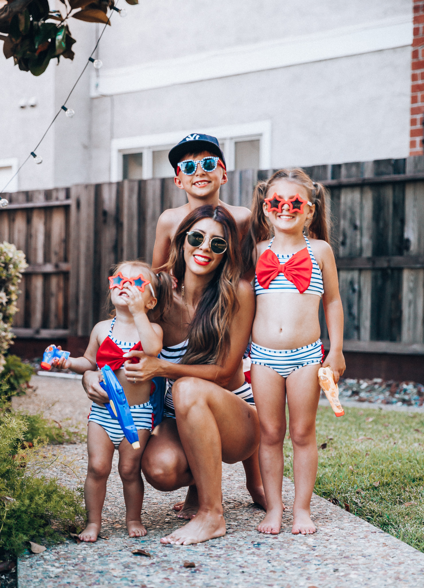 Water Gun Fights + Cute Summer Swimsuits for the Family by popular San Francisco fashion blog, The Girl in the Yellow Dress: image of a woman, two young girls and young boy standing outside holding water guns and wearing red, white and blue RuffleButts Little Girls Bikini 2-Piece Swimsuit with Bow and Ruffles, star and grinderPUNCH Kids American USA Flag Sunglasses, ON-FIELD COLLECTION Kids/Youth Yankees 59Fifty Fitted Navy Hat Cap, Old Navy American flag Patterned Board Shorts for Boys, navy Aerie Cutout one shoulder one piece swimsuit, and Nordstrom Ray-Ban Icons 53mm Retro Sunglasses.