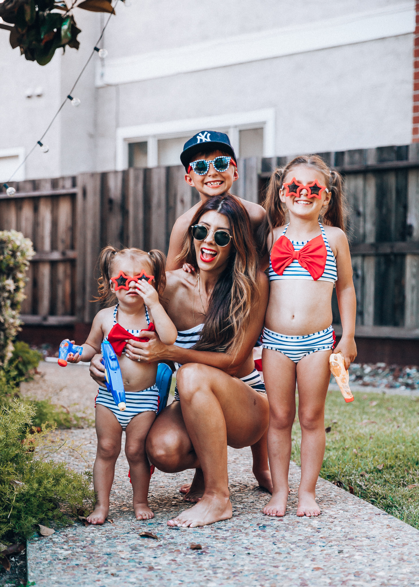 Water Gun Fights + Cute Summer Swimsuits for the Family by popular San Francisco fashion blog, The Girl in the Yellow Dress: image of a woman, two young girls and young boy standing outside holding water guns and wearing red, white and blue RuffleButts Little Girls Bikini 2-Piece Swimsuit with Bow and Ruffles, star and grinderPUNCH Kids American USA Flag Sunglasses, ON-FIELD COLLECTION Kids/Youth Yankees 59Fifty Fitted Navy Hat Cap, Old Navy American flag Patterned Board Shorts for Boys, navy Aerie Cutout one shoulder one piece swimsuit, and Nordstrom Ray-Ban Icons 53mm Retro Sunglasses.