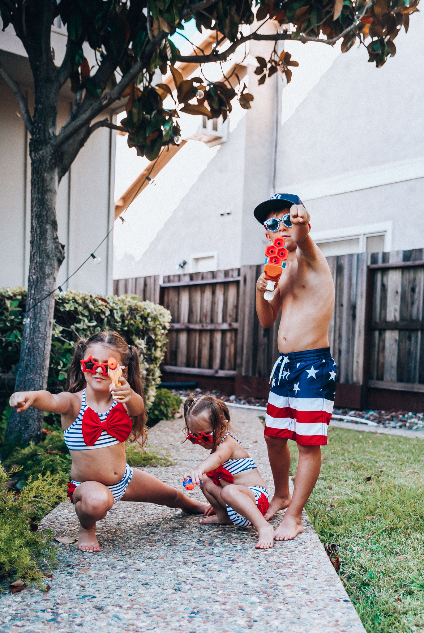 Water Gun Fights + Cute Summer Swimsuits for the Family by popular San Francisco fashion blog, The Girl in the Yellow Dress: image of two young girls and young boy standing outside holding water guns and wearing red, white and blue RuffleButts Little Girls Bikini 2-Piece Swimsuit with Bow and Ruffles, star and grinderPUNCH Kids American USA Flag Sunglasses, ON-FIELD COLLECTION Kids/Youth Yankees 59Fifty Fitted Navy Hat Cap, and Old Navy American flag Patterned Board Shorts for Boys. 