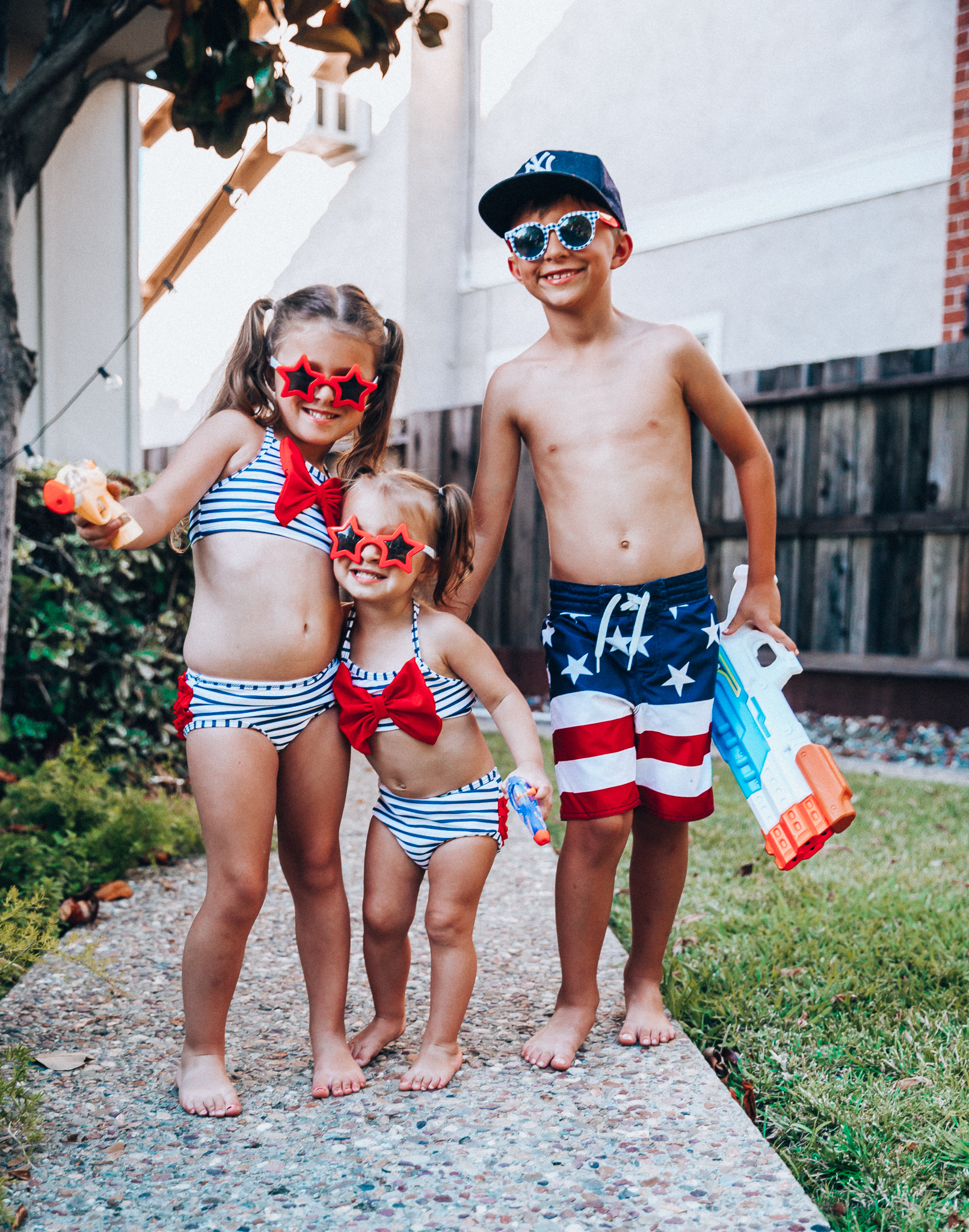 Water Gun Fights + Cute Summer Swimsuits for the Family by popular San Francisco fashion blog, The Girl in the Yellow Dress: image of two young girls and young boy standing outside holding water guns and wearing red, white and blue RuffleButts Little Girls Bikini 2-Piece Swimsuit with Bow and Ruffles, star and grinderPUNCH Kids American USA Flag Sunglasses, ON-FIELD COLLECTION Kids/Youth Yankees 59Fifty Fitted Navy Hat Cap, and Old Navy American flag Patterned Board Shorts for Boys. 