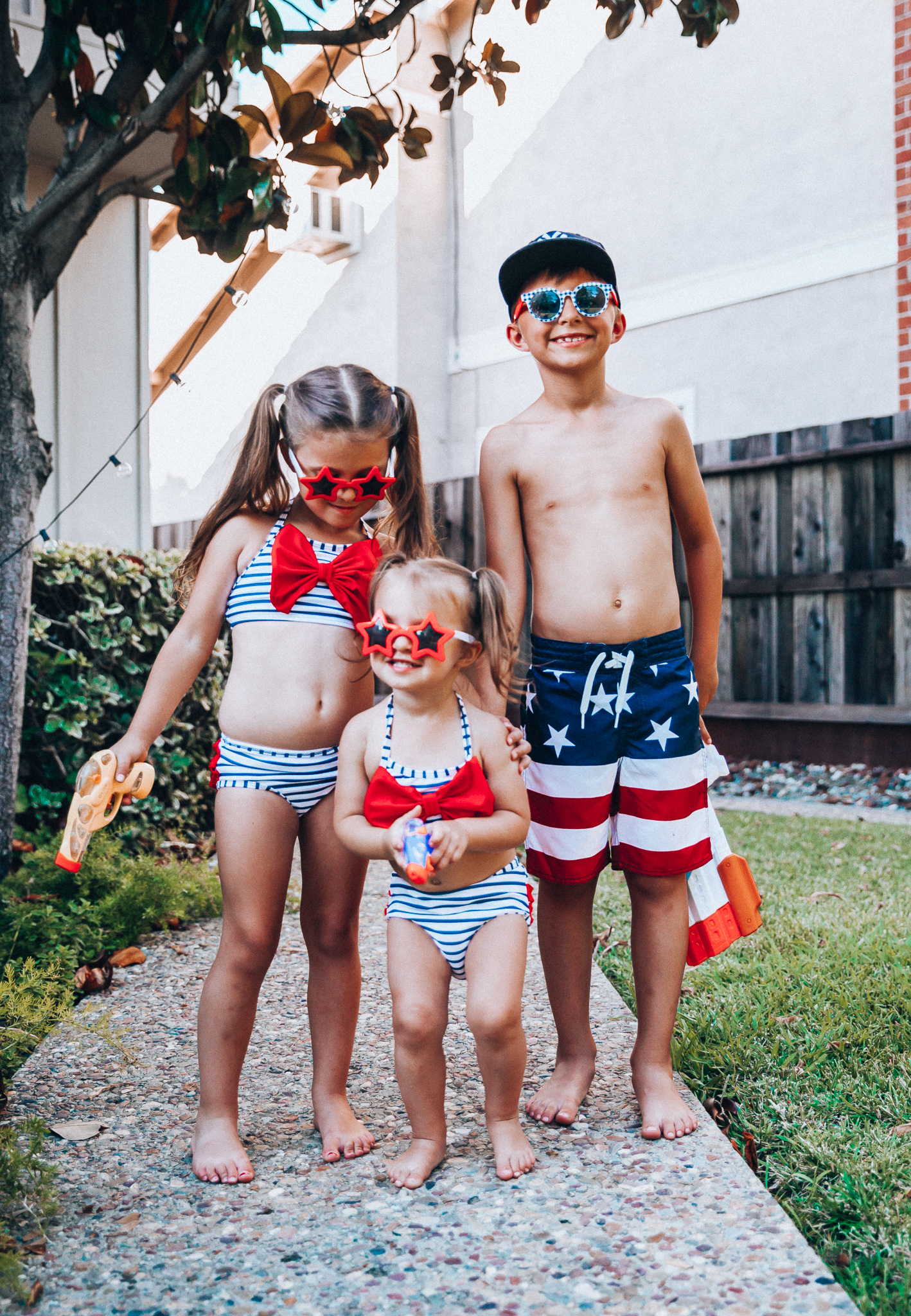 Water Gun Fights + Cute Summer Swimsuits for the Family by popular San Francisco fashion blog, The Girl in the Yellow Dress: image of two young girls and young boy standing outside holding water guns and wearing red, white and blue RuffleButts Little Girls Bikini 2-Piece Swimsuit with Bow and Ruffles, star and grinderPUNCH Kids American USA Flag Sunglasses, ON-FIELD COLLECTION Kids/Youth Yankees 59Fifty Fitted Navy Hat Cap, and Old Navy American flag Patterned Board Shorts for Boys. 