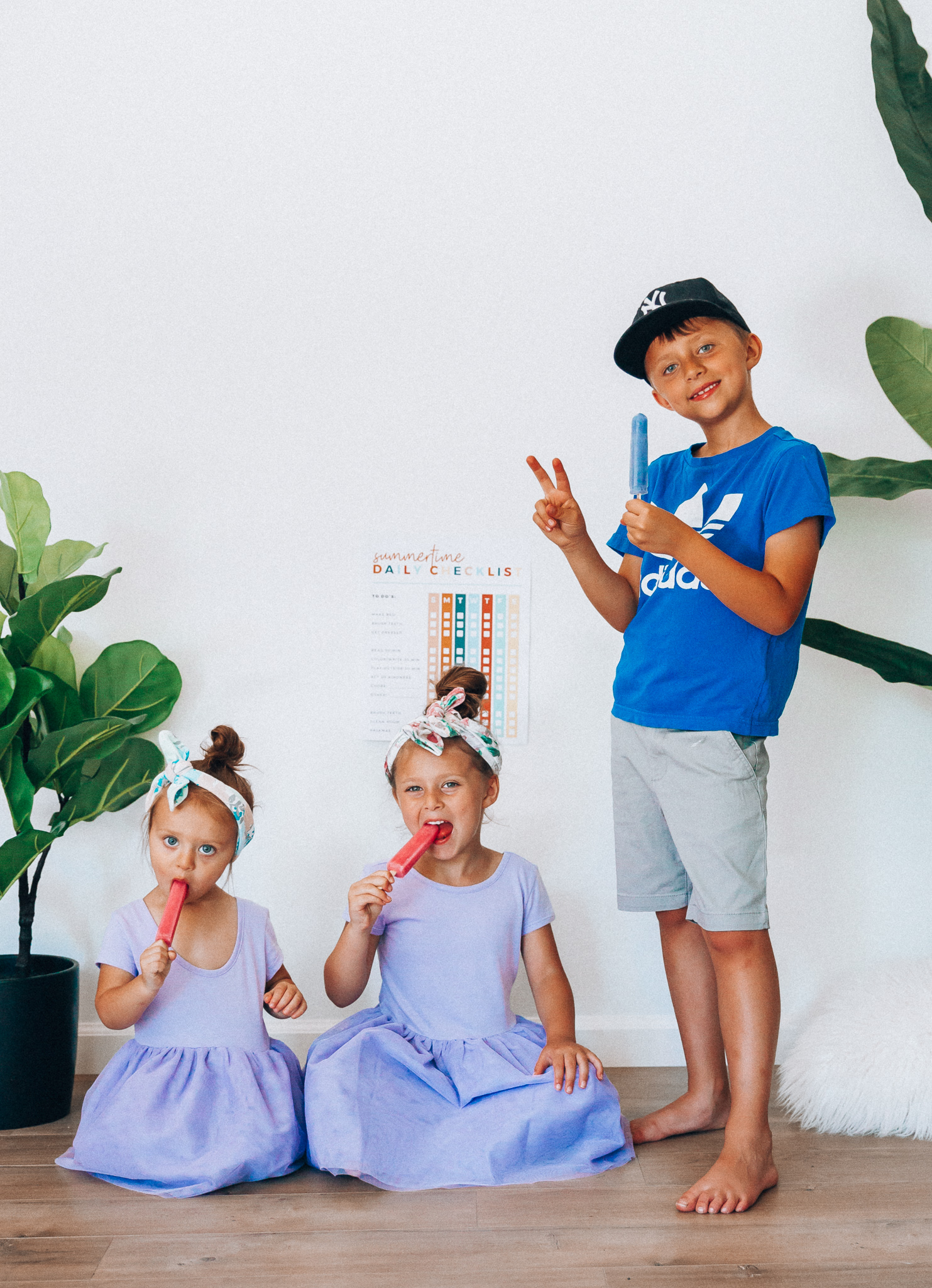 Summer Daily Checklist for Kids by popular San Francisco mom blog, The Girl in the Yellow Dress: image of two young girls and a young boy sitting in front of a daily checklist chart and eating popsicles.  The two young girls are wearing Old Navy Fit & Flare Tutu Dress for Toddler Girls, and the boy is wearing adidas Originals Baby Boys Originals Trefoil Tee, Old Navy Built-In Flex Twill Jogger Shorts for Boys, and ON-FIELD COLLECTION Kids/Youth Yankees 59Fifty Fitted Navy Hat Cap 17GM.