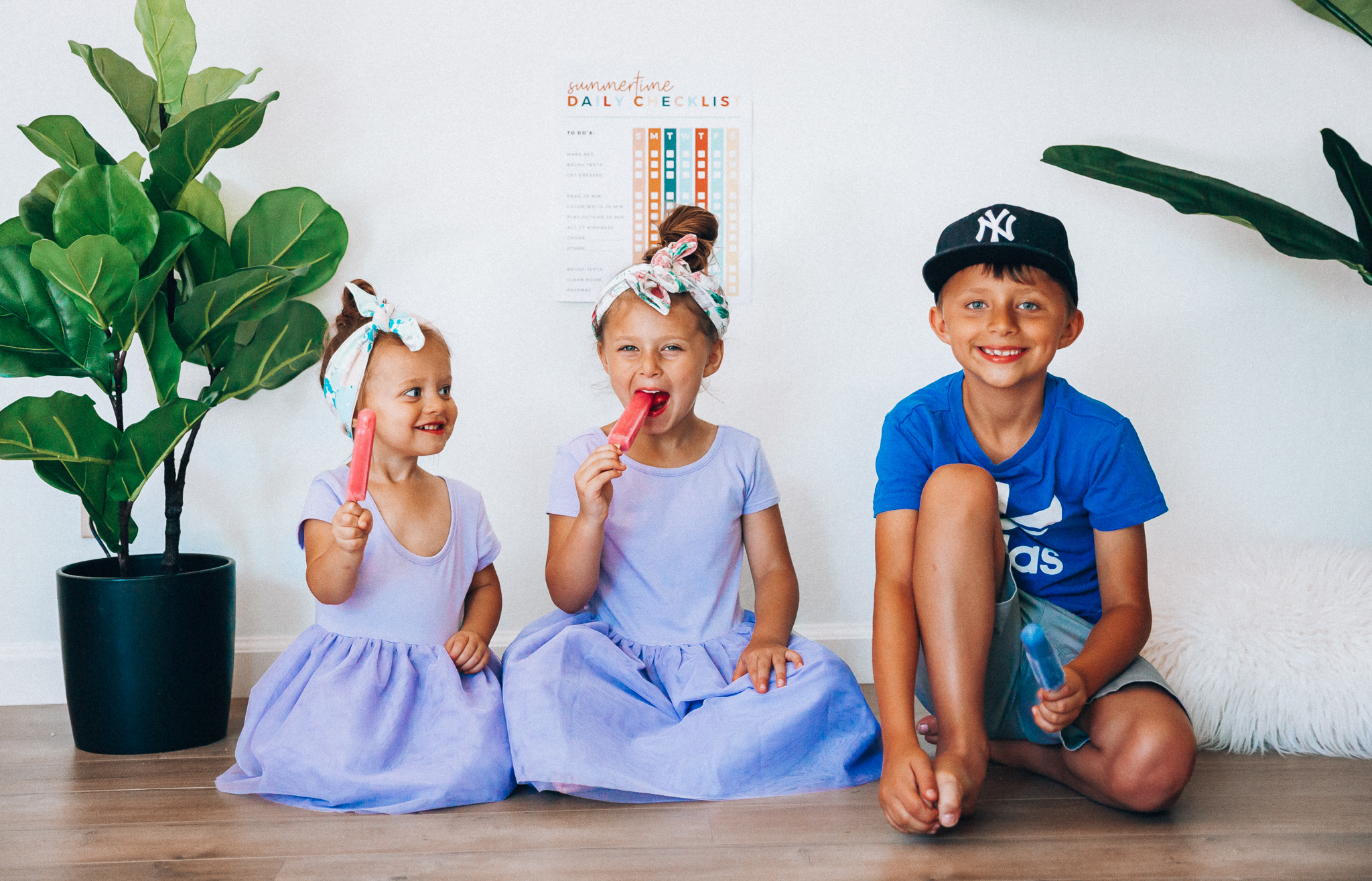 Summer Daily Checklist for Kids by popular San Francisco mom blog, The Girl in the Yellow Dress: image of two young girls and a young boy sitting in front of a daily checklist chart and eating popsicles.  The two young girls are wearing Old Navy Fit & Flare Tutu Dress for Toddler Girls, and the boy is wearing adidas Originals Baby Boys Originals Trefoil Tee, Old Navy Built-In Flex Twill Jogger Shorts for Boys, and ON-FIELD COLLECTION Kids/Youth Yankees 59Fifty Fitted Navy Hat Cap 17GM.