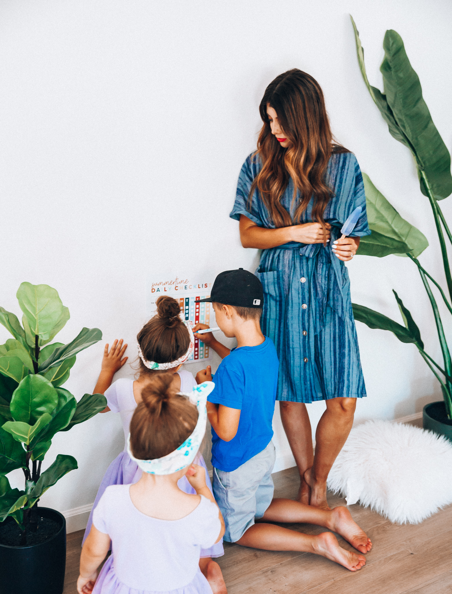 Summer Daily Checklist for Kids by popular San Francisco mom blog, The Girl in the Yellow Dress: image of woman standing with two young girls and a young boy who are marking things off on a daily checklist chart.  Mom is wearing a blue stripe dress, girls are wearing Old Navy Fit & Flare Tutu Dress for Toddler Girls, and the boy is wearing adidas Originals Baby Boys Originals Trefoil Tee, Old Navy Built-In Flex Twill Jogger Shorts for Boys, and ON-FIELD COLLECTION Kids/Youth Yankees 59Fifty Fitted Navy Hat Cap 17GM.