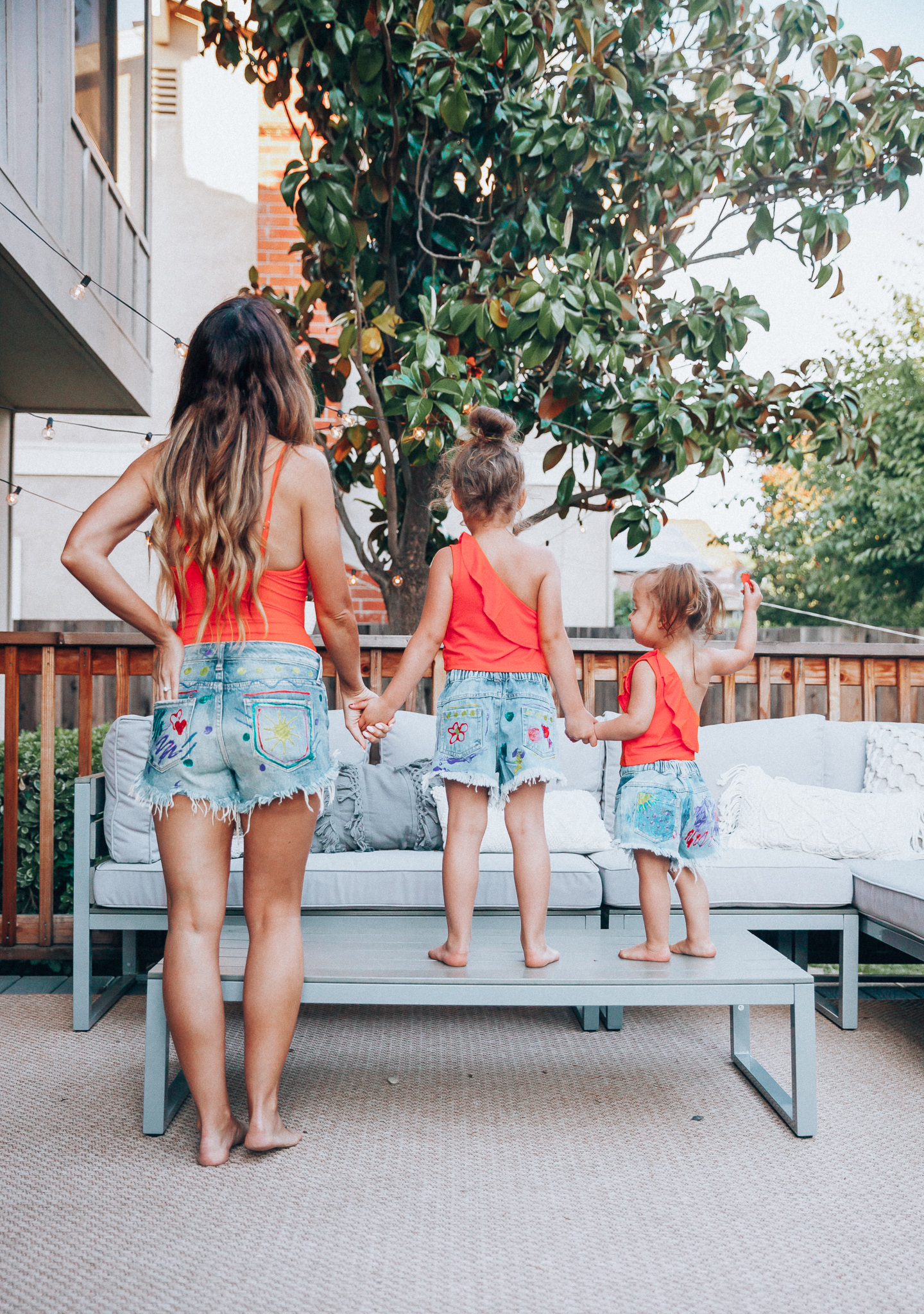 The Girl in the Yellow Dress | Latisha Springer | Fun Summer Activities for Kids by top US mom blog, The Girl in the Yellow Dress: image of mom with young girls sitting on back porch deck wearing red swimsuits and cutoff denim shorts with decorative puff paint designs.