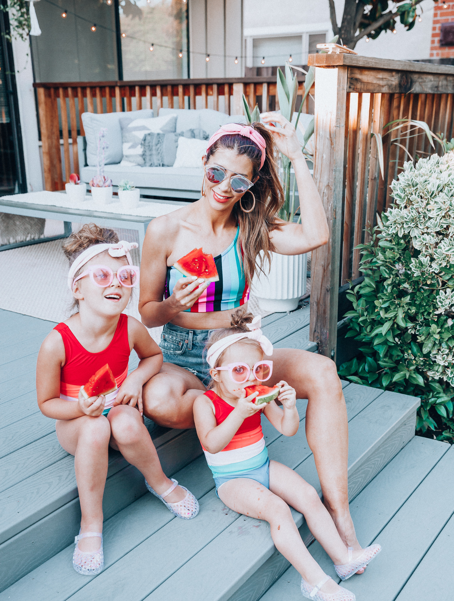 4th of July Sales by popular San Francisco fashion blog, The Girl in the Yellow Dress: image of a mom and two daughters outside on a back patio with grey patio furniture and bistro lighting. The wearing Evereve Agolde Parker Shorts, Pop Your Dream Vintage Adults Elastic Headband Bunny Ears Bow Hairband Hair Decor Accessory, MVMT Nightowl Sunglasses, and Old Navy color block one-shoulder swim top and the daughters are wearing Old Navy Color-Blocked Swimsuit for Toddler Girls, clear Basket-Weave Jelly Ballet Flats For Toddler Girls, Target Cat and Jack Girls' Cateye Sunglasses, and Pop Your Dream Vintage Adults Elastic Headband Bunny Ears Bow Hairband Hair Decor Accessory.