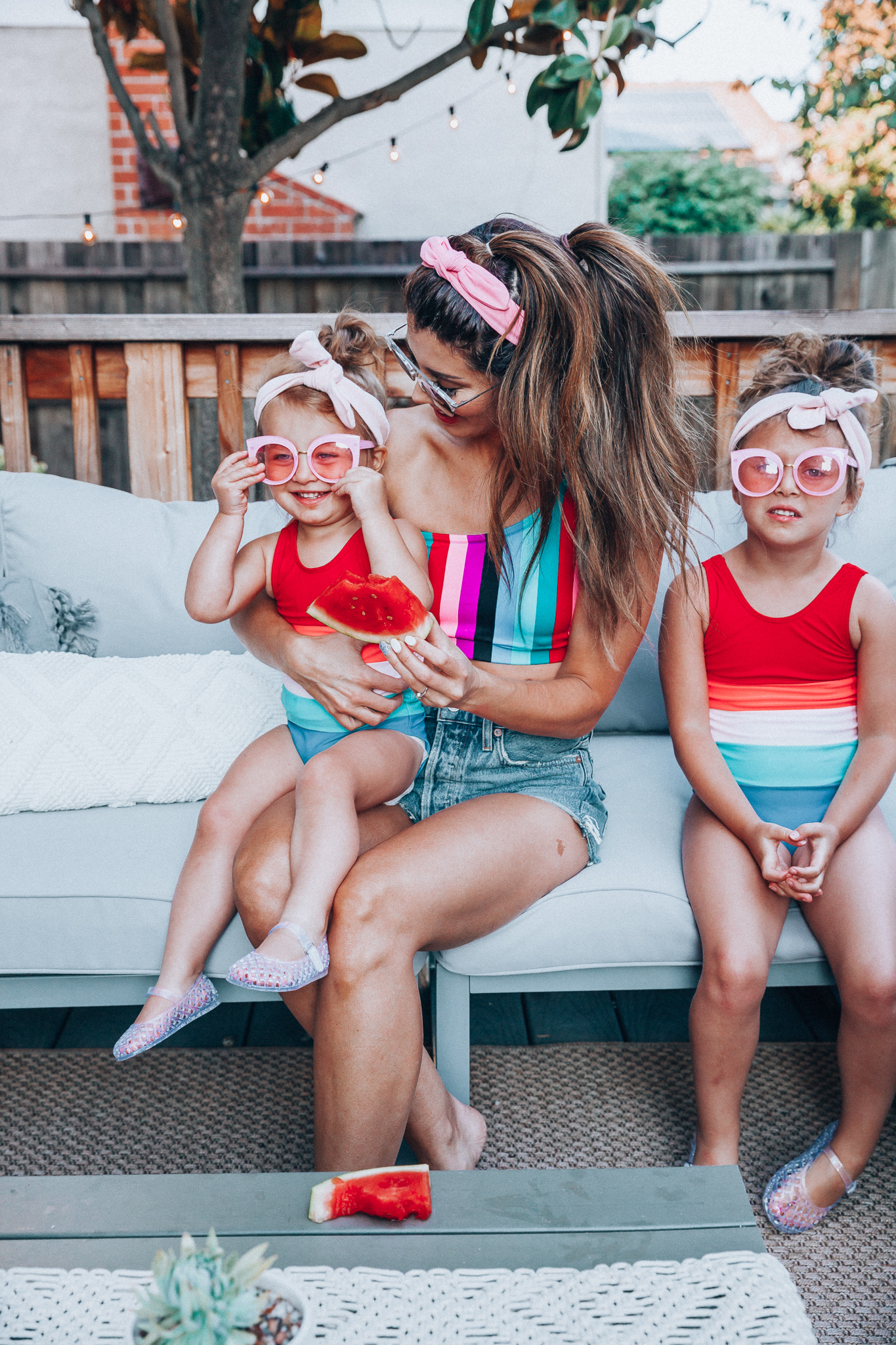 4th of July Sales by popular San Francisco fashion blog, The Girl in the Yellow Dress: image of a mom and two daughters outside on a back patio with grey patio furniture and bistro lighting. The wearing Evereve Agolde Parker Shorts, Pop Your Dream Vintage Adults Elastic Headband Bunny Ears Bow Hairband Hair Decor Accessory, MVMT Nightowl Sunglasses, and Old Navy color block one-shoulder swim top and the daughters are wearing Old Navy Color-Blocked Swimsuit for Toddler Girls, clear Basket-Weave Jelly Ballet Flats For Toddler Girls, Target Cat and Jack Girls' Cateye Sunglasses, and Pop Your Dream Vintage Adults Elastic Headband Bunny Ears Bow Hairband Hair Decor Accessory.