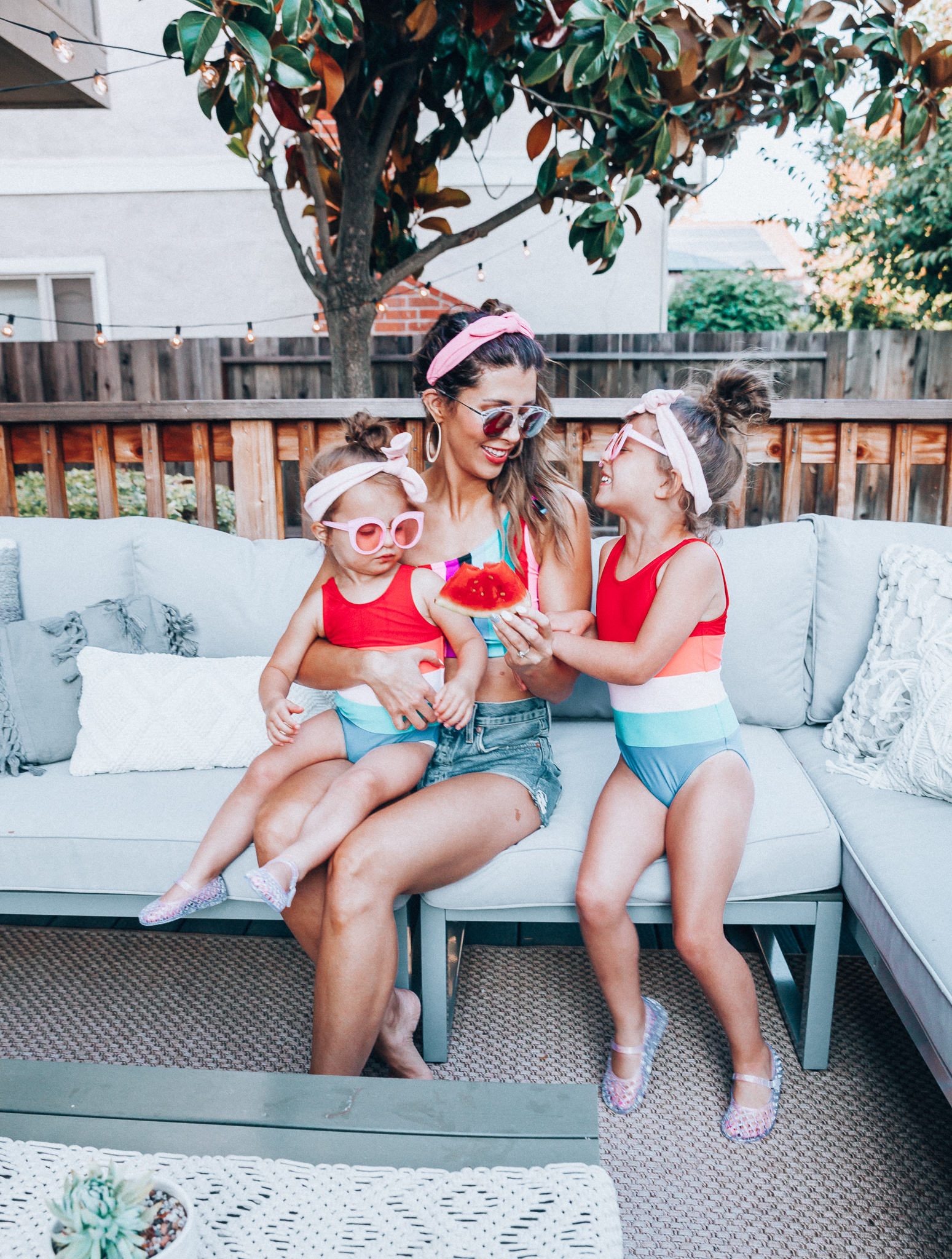 4th of July Sales by popular San Francisco fashion blog, The Girl in the Yellow Dress: image of a mom and two daughters outside on a back patio with grey patio furniture and bistro lighting. The wearing Evereve Agolde Parker Shorts, Pop Your Dream Vintage Adults Elastic Headband Bunny Ears Bow Hairband Hair Decor Accessory, MVMT Nightowl Sunglasses, and Old Navy color block one-shoulder swim top and the daughters are wearing Old Navy Color-Blocked Swimsuit for Toddler Girls, clear Basket-Weave Jelly Ballet Flats For Toddler Girls, Target Cat and Jack Girls' Cateye Sunglasses, and Pop Your Dream Vintage Adults Elastic Headband Bunny Ears Bow Hairband Hair Decor Accessory.