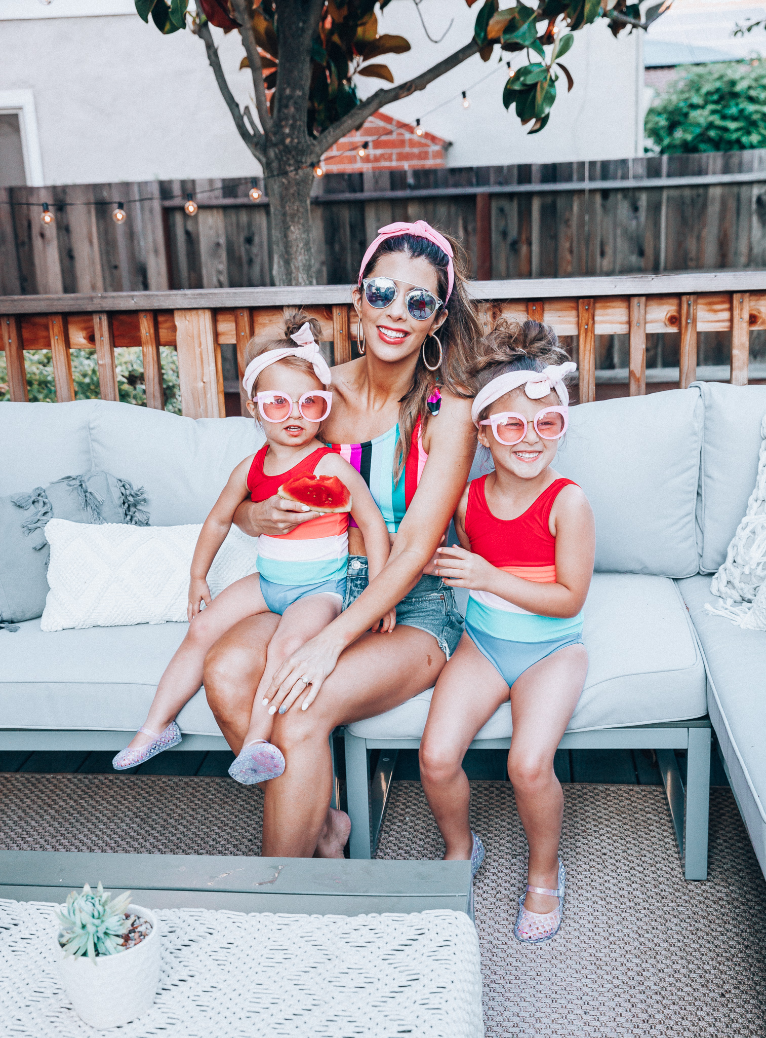 4th of July Sales by popular San Francisco fashion blog, The Girl in the Yellow Dress: image of a mom and two daughters outside on a back patio with grey patio furniture and bistro lighting. The wearing Evereve Agolde Parker Shorts, Pop Your Dream Vintage Adults Elastic Headband Bunny Ears Bow Hairband Hair Decor Accessory, MVMT Nightowl Sunglasses, and Old Navy color block one-shoulder swim top and the daughters are wearing Old Navy Color-Blocked Swimsuit for Toddler Girls, clear Basket-Weave Jelly Ballet Flats For Toddler Girls, Target Cat and Jack Girls' Cateye Sunglasses, and Pop Your Dream Vintage Adults Elastic Headband Bunny Ears Bow Hairband Hair Decor Accessory.