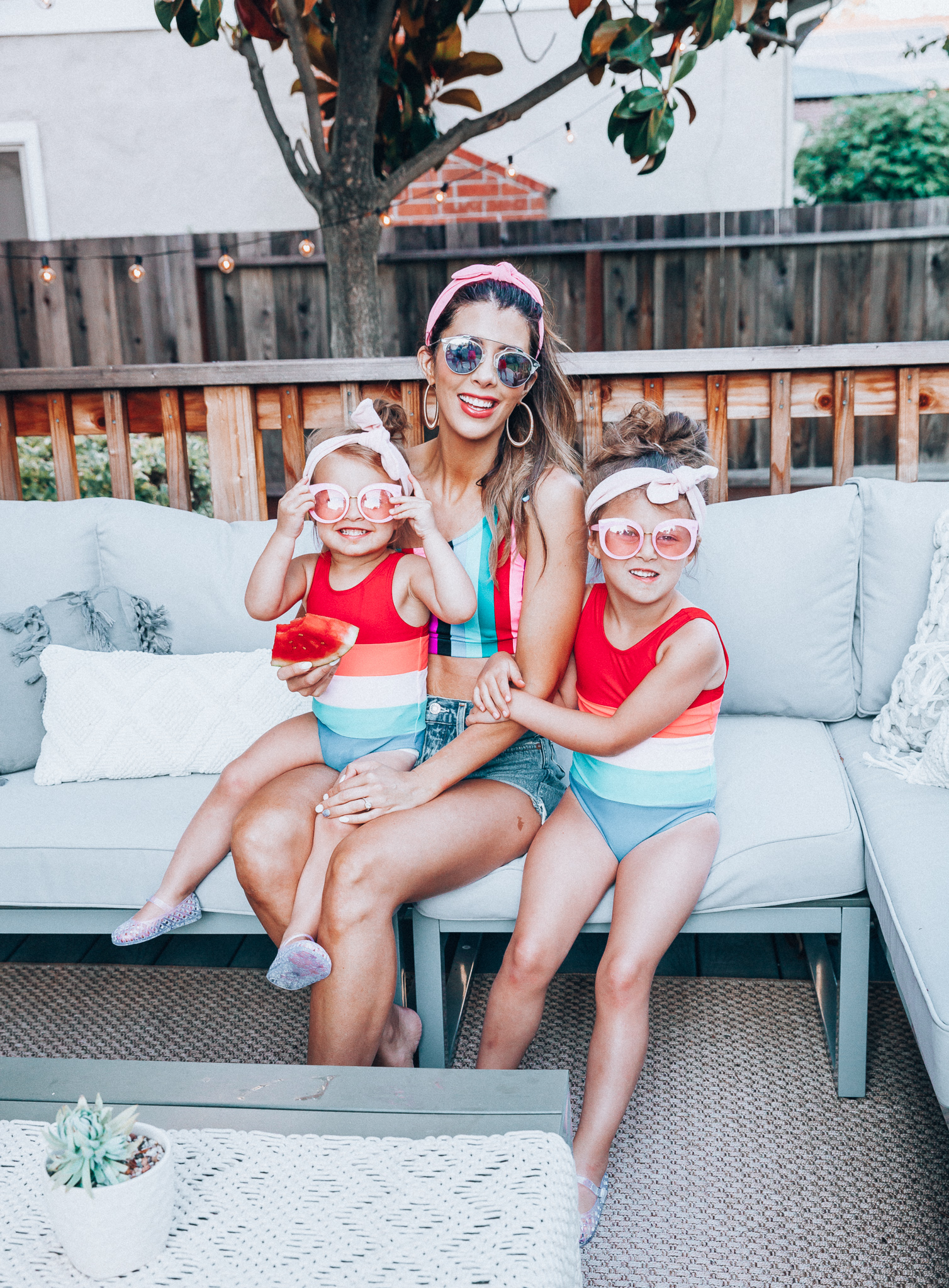 4th of July Sales by popular San Francisco fashion blog, The Girl in the Yellow Dress: image of a mom and two daughters outside on a back patio with grey patio furniture and bistro lighting. The wearing Evereve Agolde Parker Shorts, Pop Your Dream Vintage Adults Elastic Headband Bunny Ears Bow Hairband Hair Decor Accessory, MVMT Nightowl Sunglasses, and Old Navy color block one-shoulder swim top and the daughters are wearing Old Navy Color-Blocked Swimsuit for Toddler Girls, clear Basket-Weave Jelly Ballet Flats For Toddler Girls, Target Cat and Jack Girls' Cateye Sunglasses, and Pop Your Dream Vintage Adults Elastic Headband Bunny Ears Bow Hairband Hair Decor Accessory.