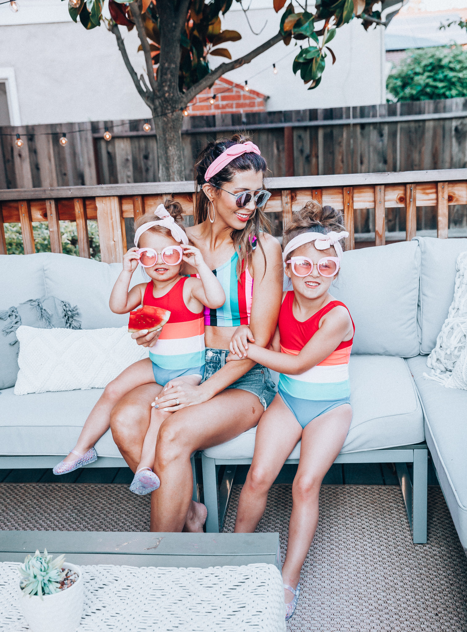 4th of July Sales by popular San Francisco fashion blog, The Girl in the Yellow Dress: image of a mom and two daughters outside on a back patio with grey patio furniture and bistro lighting. The wearing Evereve Agolde Parker Shorts, Pop Your Dream Vintage Adults Elastic Headband Bunny Ears Bow Hairband Hair Decor Accessory, MVMT Nightowl Sunglasses, and Old Navy color block one-shoulder swim top and the daughters are wearing Old Navy Color-Blocked Swimsuit for Toddler Girls, clear Basket-Weave Jelly Ballet Flats For Toddler Girls, Target Cat and Jack Girls' Cateye Sunglasses, and Pop Your Dream Vintage Adults Elastic Headband Bunny Ears Bow Hairband Hair Decor Accessory.