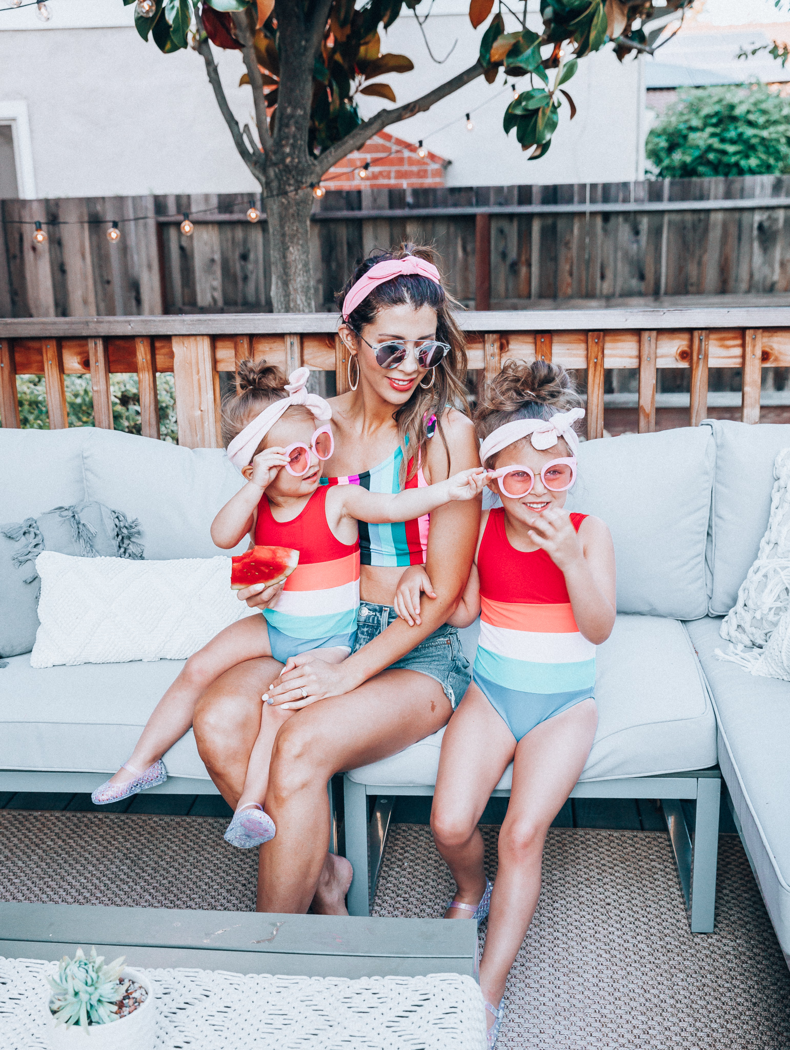 4th of July Sales by popular San Francisco fashion blog, The Girl in the Yellow Dress: image of a mom and two daughters outside on a back patio with grey patio furniture and bistro lighting. The wearing Evereve Agolde Parker Shorts, Pop Your Dream Vintage Adults Elastic Headband Bunny Ears Bow Hairband Hair Decor Accessory, MVMT Nightowl Sunglasses, and Old Navy color block one-shoulder swim top and the daughters are wearing Old Navy Color-Blocked Swimsuit for Toddler Girls, clear Basket-Weave Jelly Ballet Flats For Toddler Girls, Target Cat and Jack Girls' Cateye Sunglasses, and Pop Your Dream Vintage Adults Elastic Headband Bunny Ears Bow Hairband Hair Decor Accessory.