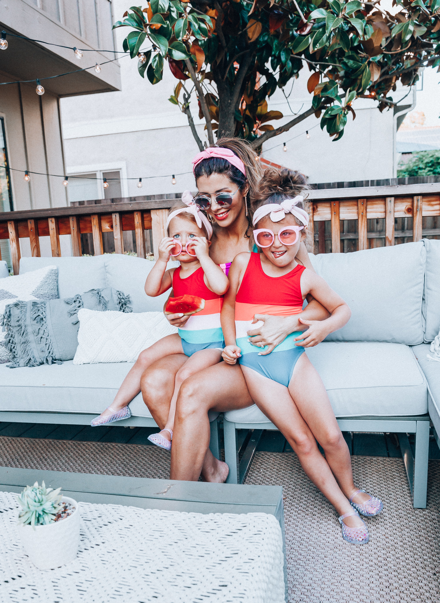 4th of July Sales by popular San Francisco fashion blog, The Girl in the Yellow Dress: image of a mom and two daughters outside on a back patio with grey patio furniture and bistro lighting. The wearing Evereve Agolde Parker Shorts, Pop Your Dream Vintage Adults Elastic Headband Bunny Ears Bow Hairband Hair Decor Accessory, MVMT Nightowl Sunglasses, and Old Navy color block one-shoulder swim top and the daughters are wearing Old Navy Color-Blocked Swimsuit for Toddler Girls, clear Basket-Weave Jelly Ballet Flats For Toddler Girls, Target Cat and Jack Girls' Cateye Sunglasses, and Pop Your Dream Vintage Adults Elastic Headband Bunny Ears Bow Hairband Hair Decor Accessory.