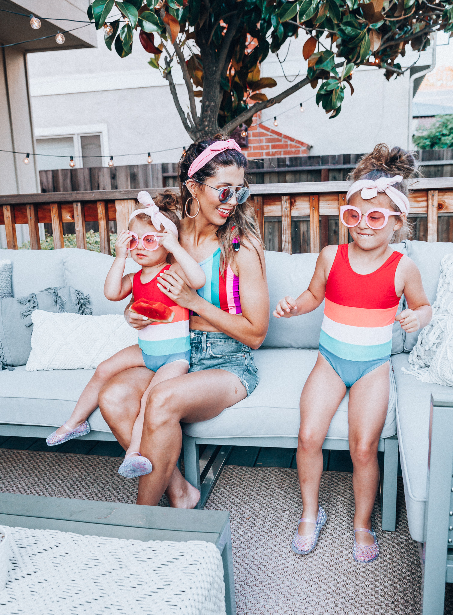 4th of July Sales by popular San Francisco fashion blog, The Girl in the Yellow Dress: image of a mom and two daughters outside on a back patio with grey patio furniture and bistro lighting. The wearing Evereve Agolde Parker Shorts, Pop Your Dream Vintage Adults Elastic Headband Bunny Ears Bow Hairband Hair Decor Accessory, MVMT Nightowl Sunglasses, and Old Navy color block one-shoulder swim top and the daughters are wearing Old Navy Color-Blocked Swimsuit for Toddler Girls, clear Basket-Weave Jelly Ballet Flats For Toddler Girls, Target Cat and Jack Girls' Cateye Sunglasses, and Pop Your Dream Vintage Adults Elastic Headband Bunny Ears Bow Hairband Hair Decor Accessory.