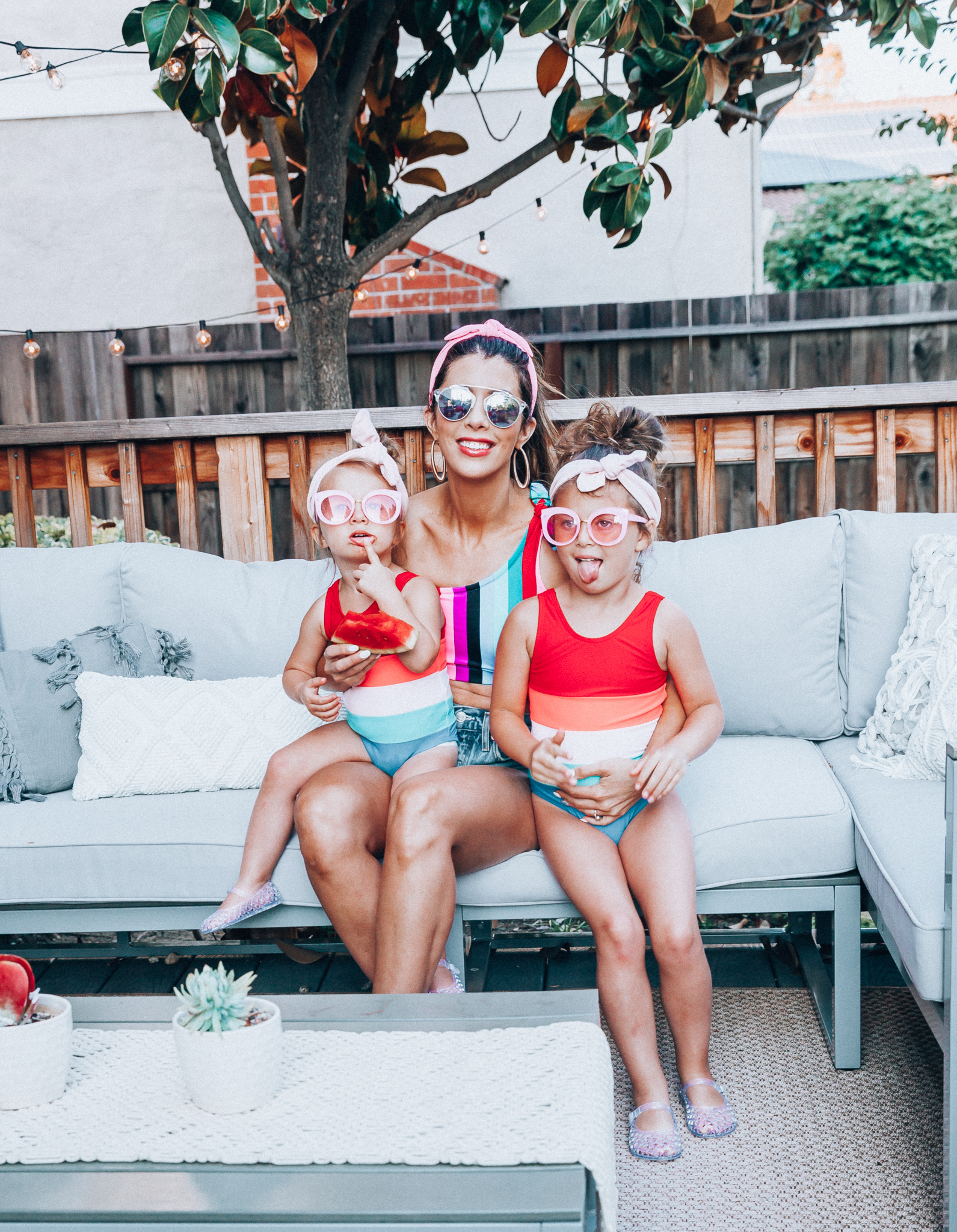 4th of July Sales by popular San Francisco fashion blog, The Girl in the Yellow Dress: image of a mom and two daughters outside on a back patio with grey patio furniture and bistro lighting. The wearing Evereve Agolde Parker Shorts, Pop Your Dream Vintage Adults Elastic Headband Bunny Ears Bow Hairband Hair Decor Accessory, MVMT Nightowl Sunglasses, and Old Navy color block one-shoulder swim top and the daughters are wearing Old Navy Color-Blocked Swimsuit for Toddler Girls, clear Basket-Weave Jelly Ballet Flats For Toddler Girls, Target Cat and Jack Girls' Cateye Sunglasses, and Pop Your Dream Vintage Adults Elastic Headband Bunny Ears Bow Hairband Hair Decor Accessory.