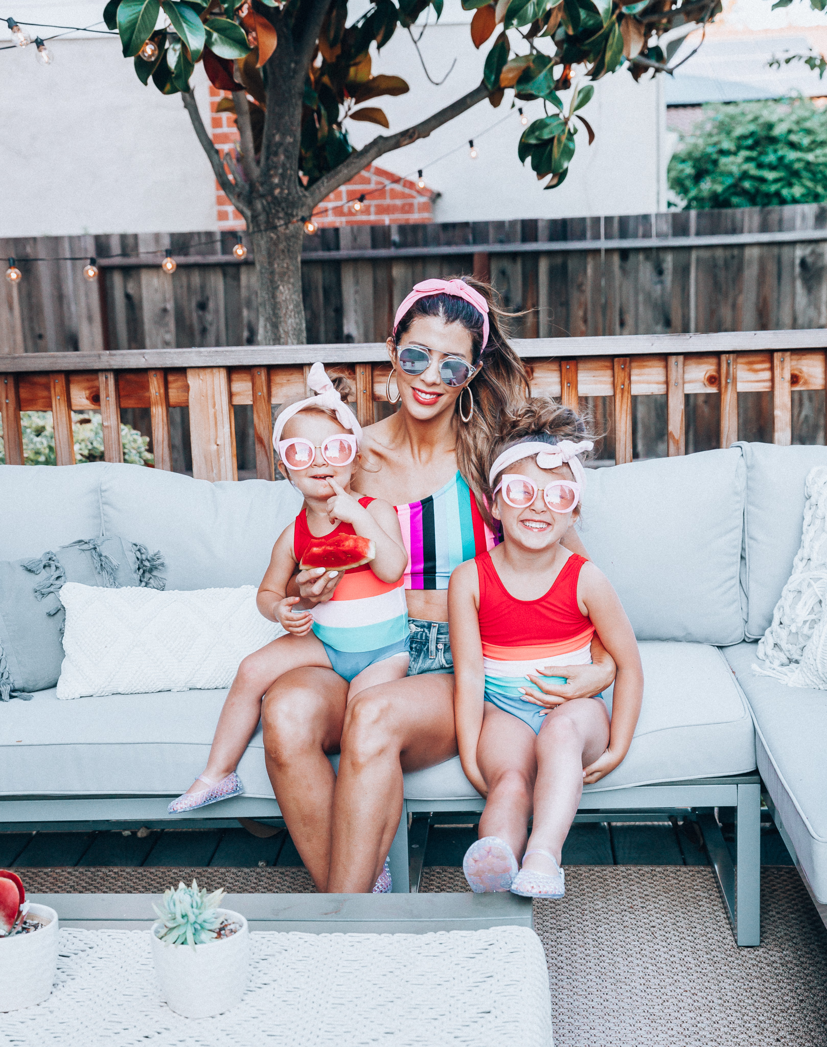 4th of July Sales by popular San Francisco fashion blog, The Girl in the Yellow Dress: image of a mom and two daughters outside on a back patio with grey patio furniture and bistro lighting. The wearing Evereve Agolde Parker Shorts, Pop Your Dream Vintage Adults Elastic Headband Bunny Ears Bow Hairband Hair Decor Accessory, MVMT Nightowl Sunglasses, and Old Navy color block one-shoulder swim top and the daughters are wearing Old Navy Color-Blocked Swimsuit for Toddler Girls, clear Basket-Weave Jelly Ballet Flats For Toddler Girls, Target Cat and Jack Girls' Cateye Sunglasses, and Pop Your Dream Vintage Adults Elastic Headband Bunny Ears Bow Hairband Hair Decor Accessory.