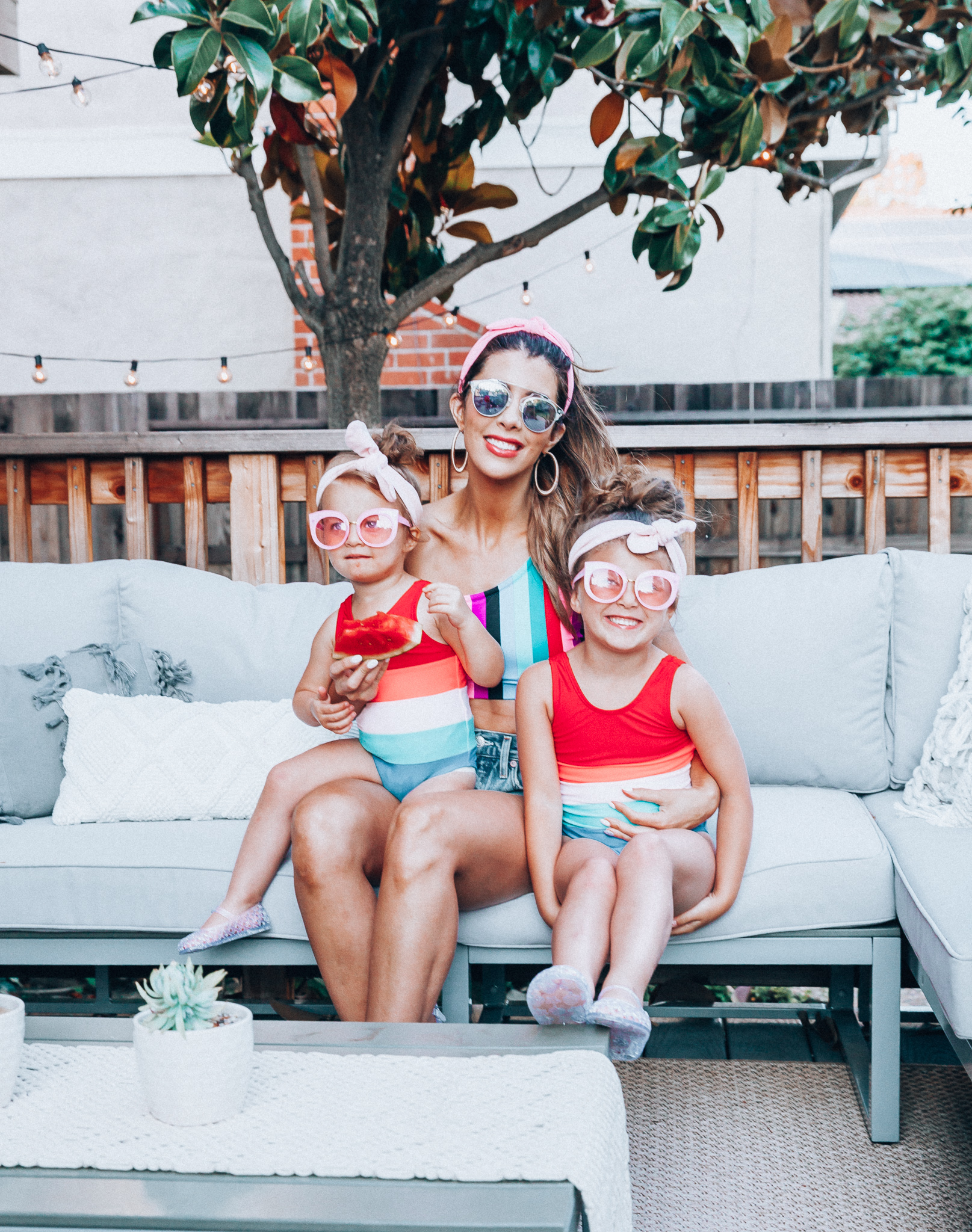 4th of July Sales by popular San Francisco fashion blog, The Girl in the Yellow Dress: image of a mom and two daughters outside on a back patio with grey patio furniture and bistro lighting. The wearing Evereve Agolde Parker Shorts, Pop Your Dream Vintage Adults Elastic Headband Bunny Ears Bow Hairband Hair Decor Accessory, MVMT Nightowl Sunglasses, and Old Navy color block one-shoulder swim top and the daughters are wearing Old Navy Color-Blocked Swimsuit for Toddler Girls, clear Basket-Weave Jelly Ballet Flats For Toddler Girls, Target Cat and Jack Girls' Cateye Sunglasses, and Pop Your Dream Vintage Adults Elastic Headband Bunny Ears Bow Hairband Hair Decor Accessory.