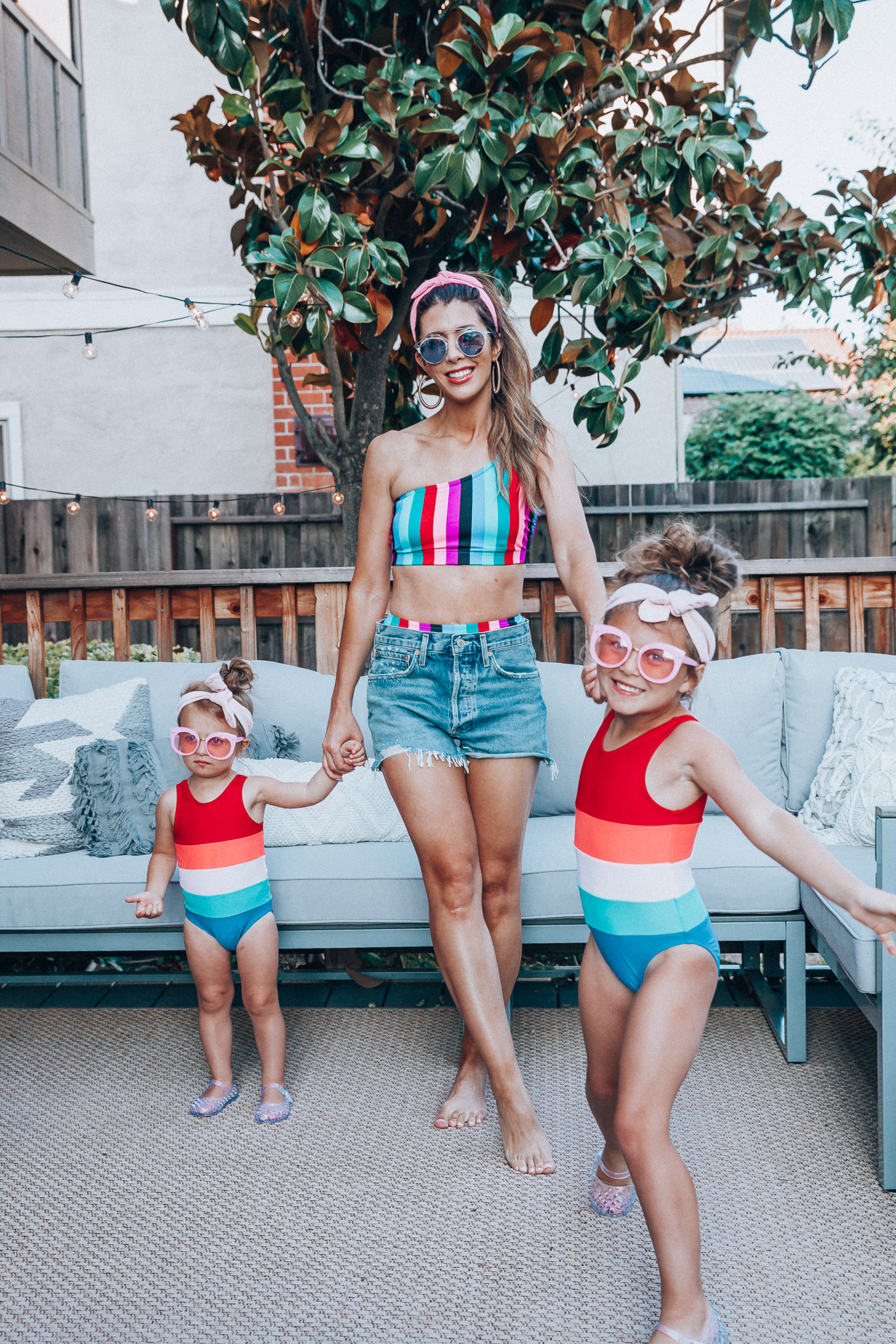 4th of July Sales by popular San Francisco fashion blog, The Girl in the Yellow Dress: image of a mom and two daughters outside on a back patio with grey patio furniture and bistro lighting. The wearing Evereve Agolde Parker Shorts, Pop Your Dream Vintage Adults Elastic Headband Bunny Ears Bow Hairband Hair Decor Accessory, MVMT Nightowl Sunglasses, and Old Navy color block one-shoulder swim top and the daughters are wearing Old Navy Color-Blocked Swimsuit for Toddler Girls, clear Basket-Weave Jelly Ballet Flats For Toddler Girls, Target Cat and Jack Girls' Cateye Sunglasses, and Pop Your Dream Vintage Adults Elastic Headband Bunny Ears Bow Hairband Hair Decor Accessory.