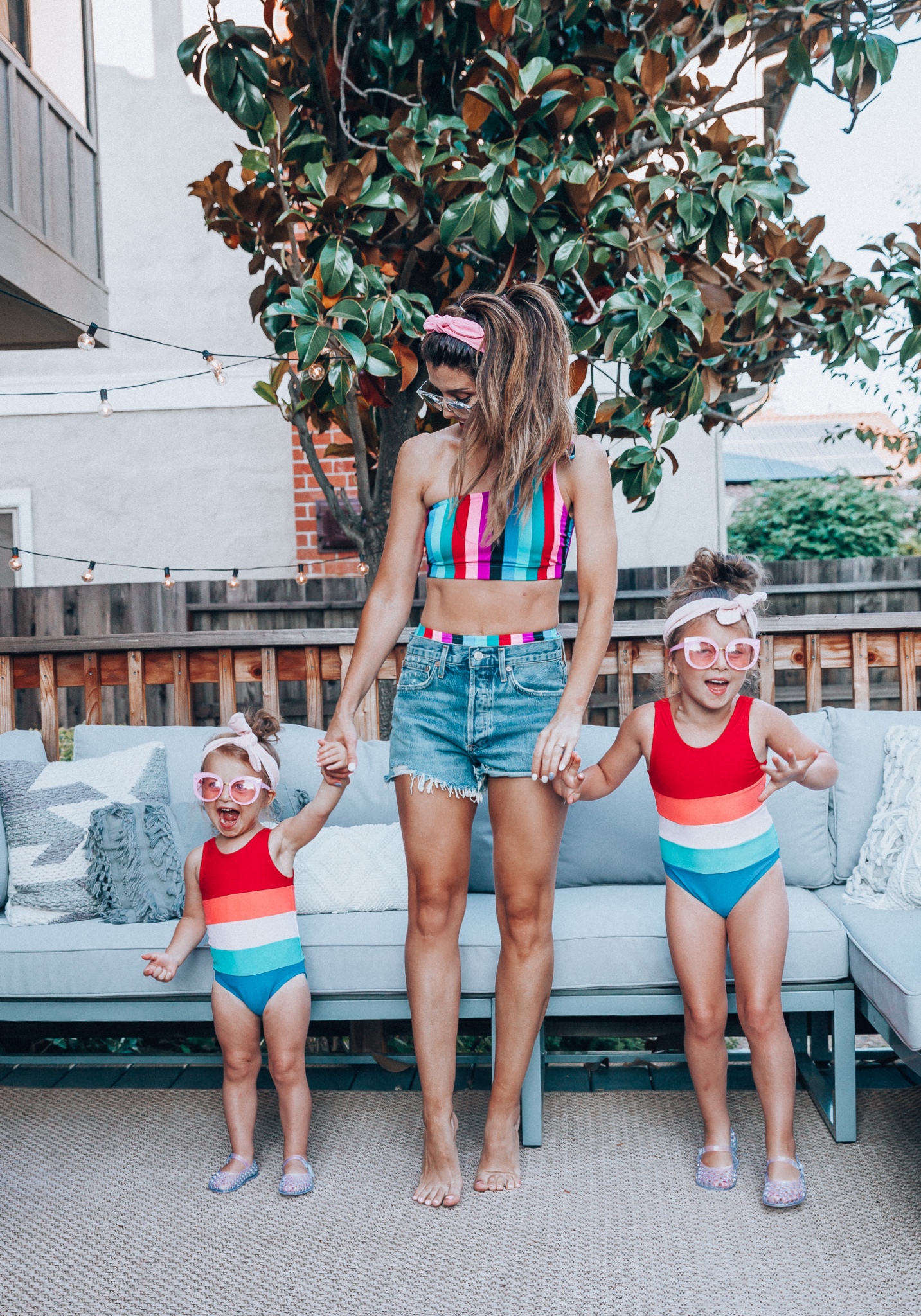 4th of July Sales by popular San Francisco fashion blog, The Girl in the Yellow Dress: image of a mom and two daughters outside on a back patio with grey patio furniture and bistro lighting. The wearing Evereve Agolde Parker Shorts, Pop Your Dream Vintage Adults Elastic Headband Bunny Ears Bow Hairband Hair Decor Accessory, MVMT Nightowl Sunglasses, and Old Navy color block one-shoulder swim top and the daughters are wearing Old Navy Color-Blocked Swimsuit for Toddler Girls, clear Basket-Weave Jelly Ballet Flats For Toddler Girls, Target Cat and Jack Girls' Cateye Sunglasses, and Pop Your Dream Vintage Adults Elastic Headband Bunny Ears Bow Hairband Hair Decor Accessory.