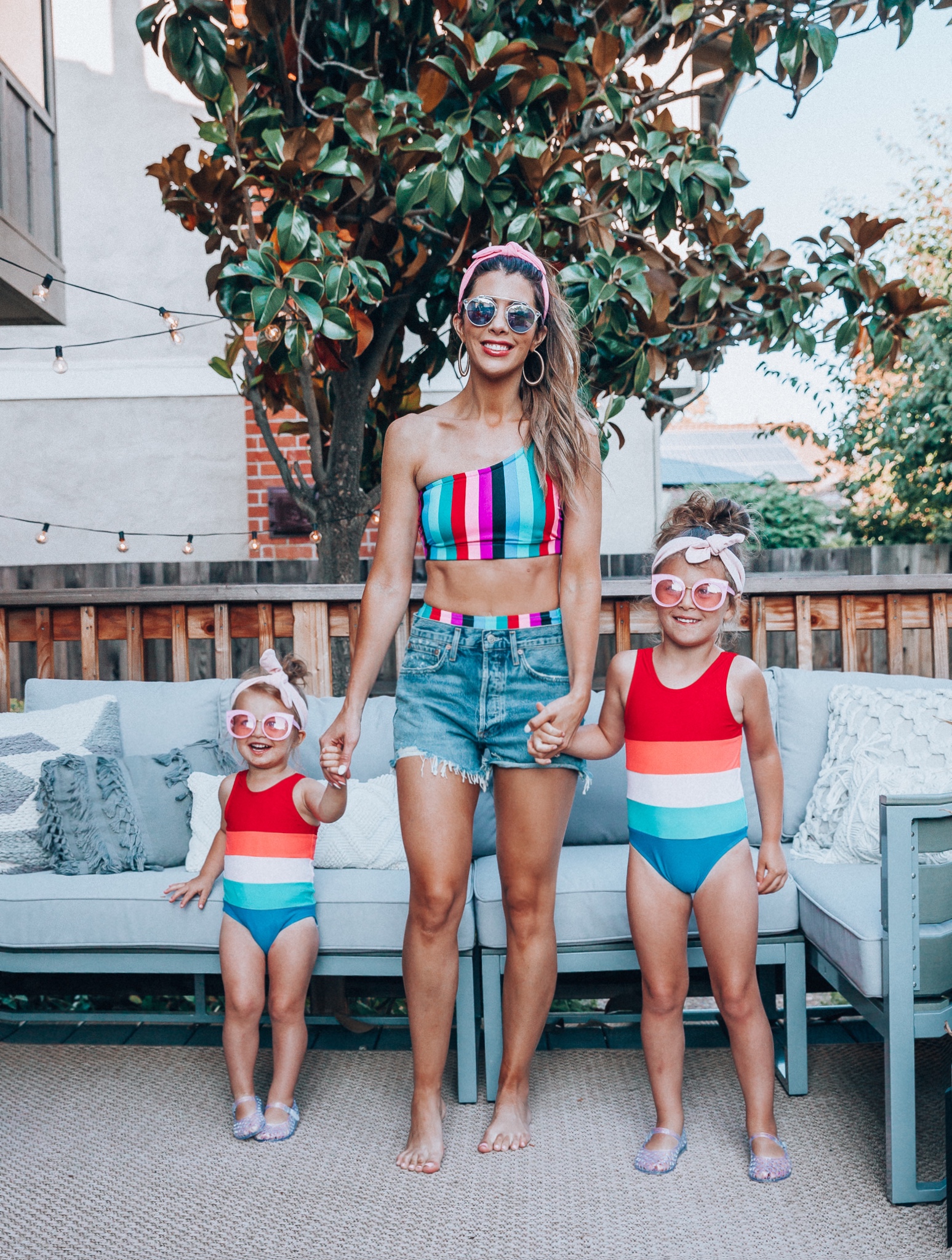 4th of July Sales by popular San Francisco fashion blog, The Girl in the Yellow Dress: image of a mom and two daughters outside on a back patio with grey patio furniture and bistro lighting. The wearing Evereve Agolde Parker Shorts, Pop Your Dream Vintage Adults Elastic Headband Bunny Ears Bow Hairband Hair Decor Accessory, MVMT Nightowl Sunglasses, and Old Navy color block one-shoulder swim top and the daughters are wearing Old Navy Color-Blocked Swimsuit for Toddler Girls, clear Basket-Weave Jelly Ballet Flats For Toddler Girls, Target Cat and Jack Girls' Cateye Sunglasses, and Pop Your Dream Vintage Adults Elastic Headband Bunny Ears Bow Hairband Hair Decor Accessory.