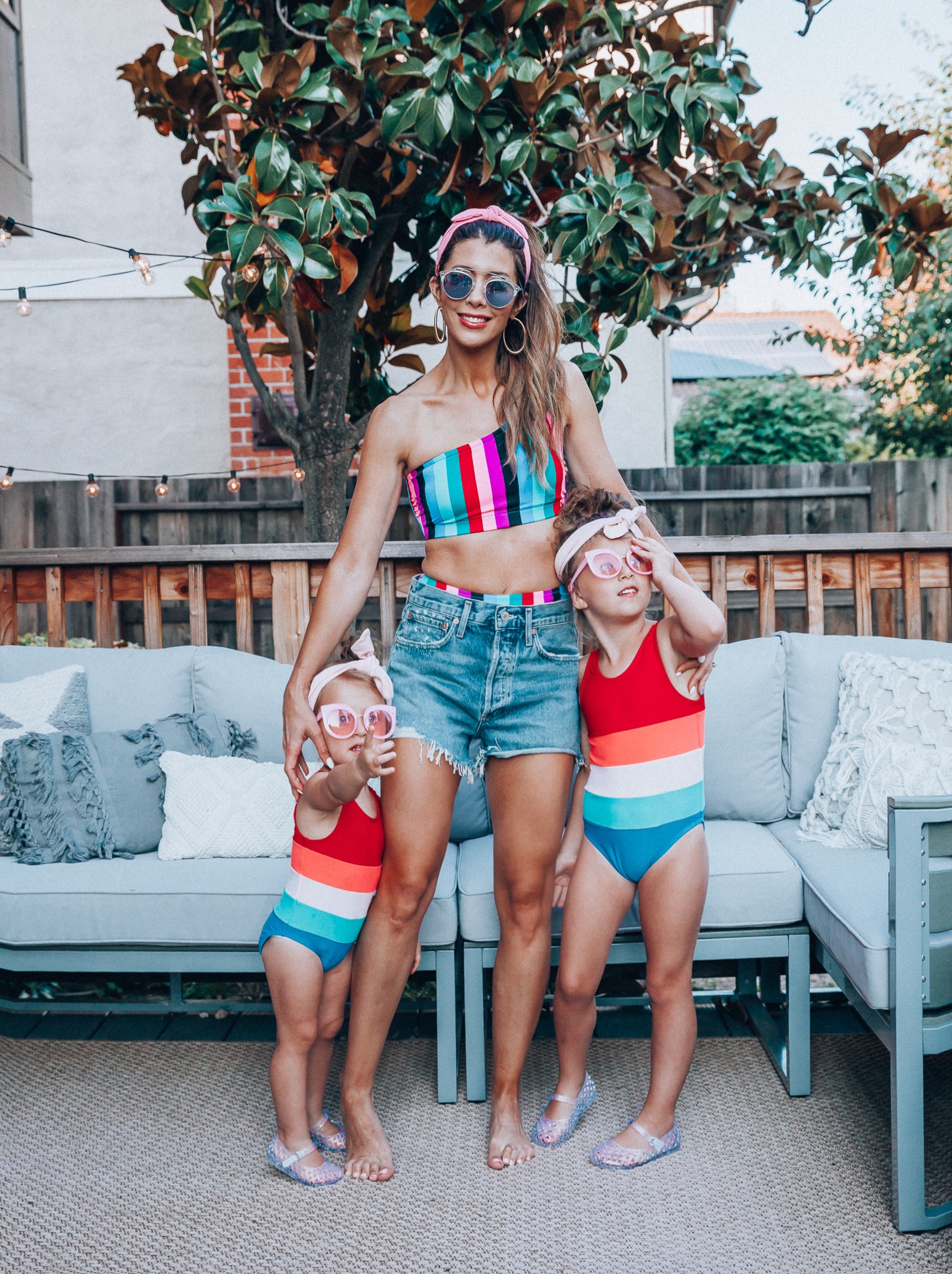 4th of July Sales by popular San Francisco fashion blog, The Girl in the Yellow Dress: image of a mom and two daughters outside on a back patio with grey patio furniture and bistro lighting. The wearing Evereve Agolde Parker Shorts, Pop Your Dream Vintage Adults Elastic Headband Bunny Ears Bow Hairband Hair Decor Accessory, MVMT Nightowl Sunglasses, and Old Navy color block one-shoulder swim top and the daughters are wearing Old Navy Color-Blocked Swimsuit for Toddler Girls, clear Basket-Weave Jelly Ballet Flats For Toddler Girls, Target Cat and Jack Girls' Cateye Sunglasses, and Pop Your Dream Vintage Adults Elastic Headband Bunny Ears Bow Hairband Hair Decor Accessory.