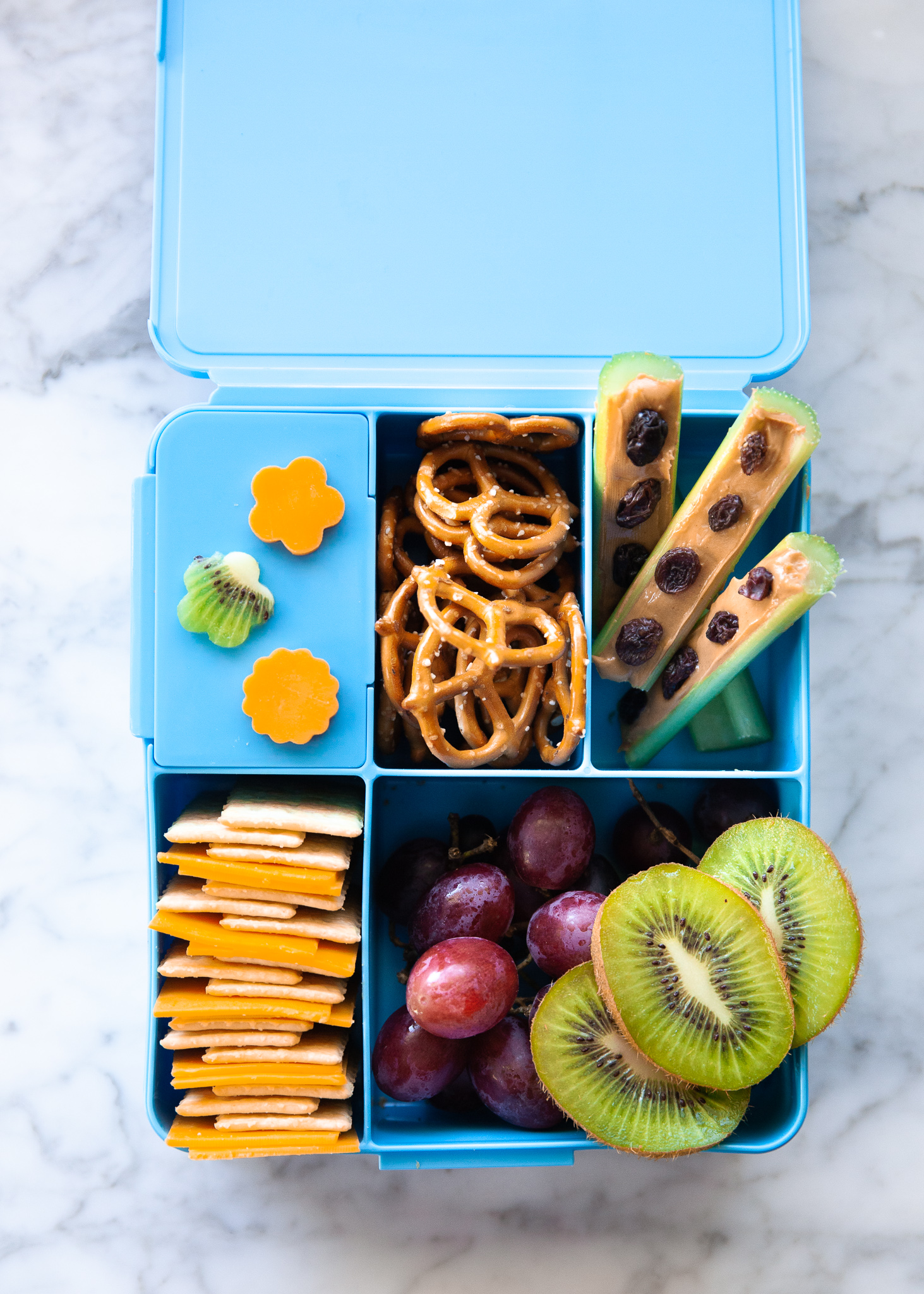 Easy Back to School Lunch Ideas! by popular San Francisco lifestyle blog, The Girl in the Yellow Dress: collage image of Pottery Barn Kids Spencer Bento Box Containers with it's compartments filled with kiwi and cheese cut into flowers shapes by a LENK Vegetable Cutter Shapes Set, pretzels, raspberries, crackers and ham slices, and celery sticks filled with peanut butter and raisins. 