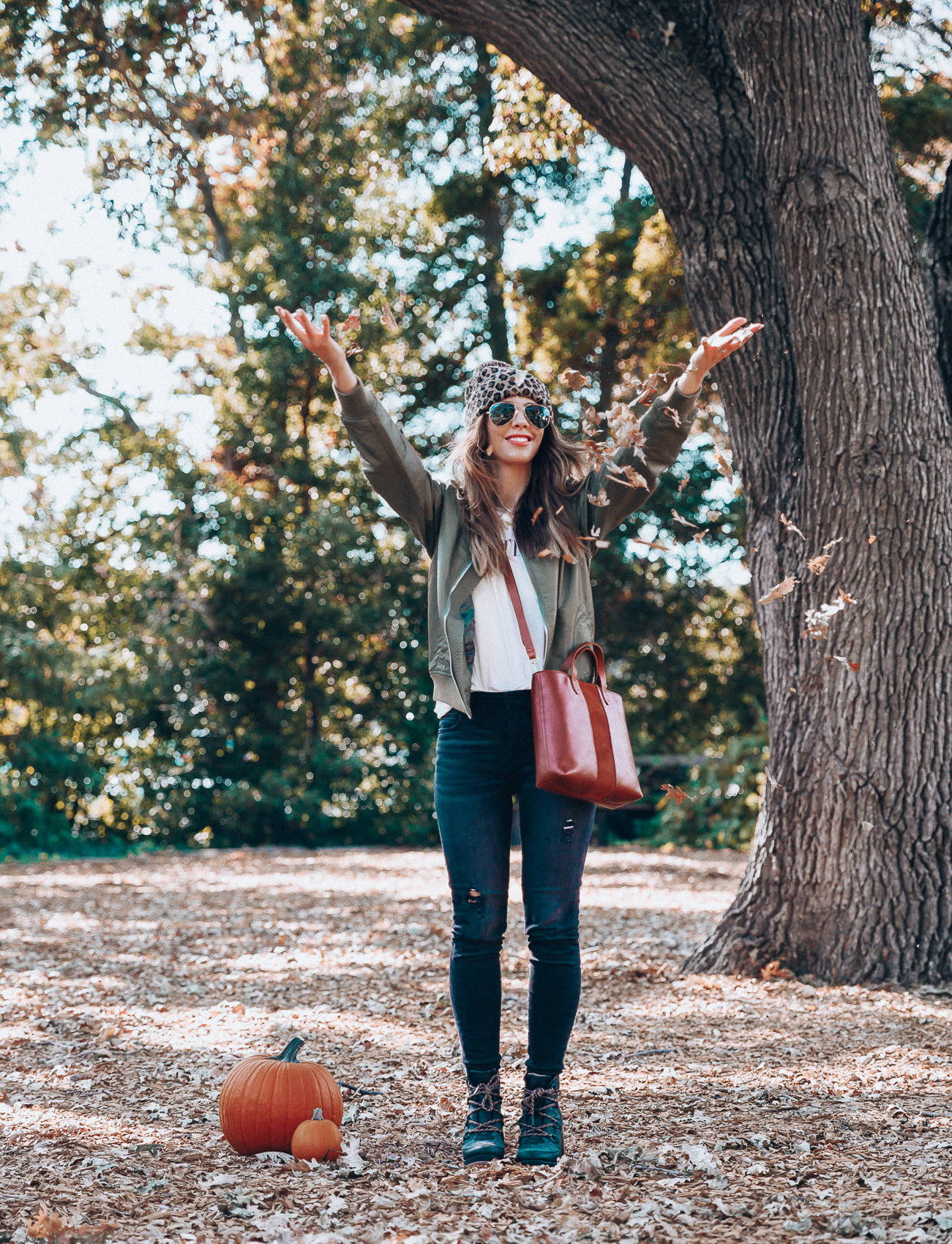 4 Fall Trends to Try by popular San Francisco fashion blog, The Girl in the Yellow Dress: image of a woman standing outside in a pile of leaves and wearing a Sole Society LEOPARD BEANIE W/ POM, EverEve Good American Bomber Jacket, Nordstrom Top Shop Jamie High Waist Ripped Hem Skinny Jeans, Evereve Sorel Blake Lace Up Bootie, Nordstrom Ray-Ban Standard Original 58mm Aviator Sunglasses, and Evereve Sol Angeles Grateful Tee.