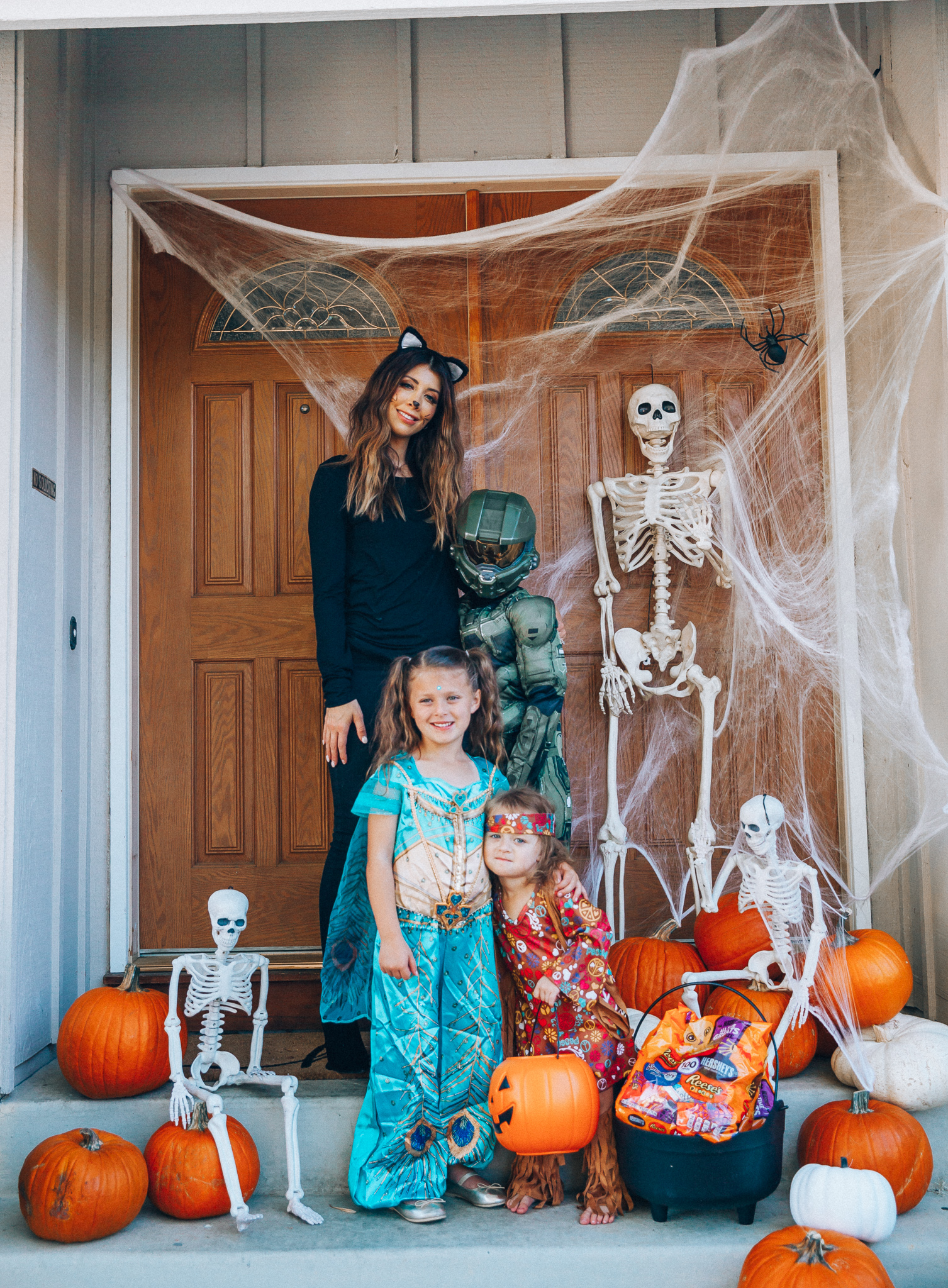 Spooky Halloween Front Porch Decor from Walmart by popular San Francisco life and style blog, The Girl in the Yellow Dress: image of a family standing on their front porch that's decorated with a Walmart 5ft Full Body Skeleton Props with Movable Joints for Halloween Party Decoration, Halloween Dutch Kettle Cauldron 14 Inch Black Plastic Party Accessory (1/Pkg) Pkg/1  Report incorrect product info or prohibited items Halloween Dutch Kettle Cauldron 14 Inch Black Plastic Party Accessory, Walmart Poseable Skeleton 20in Halloween Decoration, Pumpkins, and Walmart Stretchable Spider Web and Spider Halloween Decoration and wearing a Walmart Girl's Jasmine Teal Classic Halloween Costume, Walmart 60's Hippie with Fringe Child Halloween Costume, Walmart Halo Boys' Master Chief Classic Muscle Costume, Walmart Sequin cat ears, Walmart Jordache  Women's Super Soft Mid Rise Skinny Jean, Walmart Seven Dials  Womens Seven Dials Teatime Sock Booties, and Walmart TheLovely  Women's Long Sleeve Crew Neck Basic T-Shirt Top.