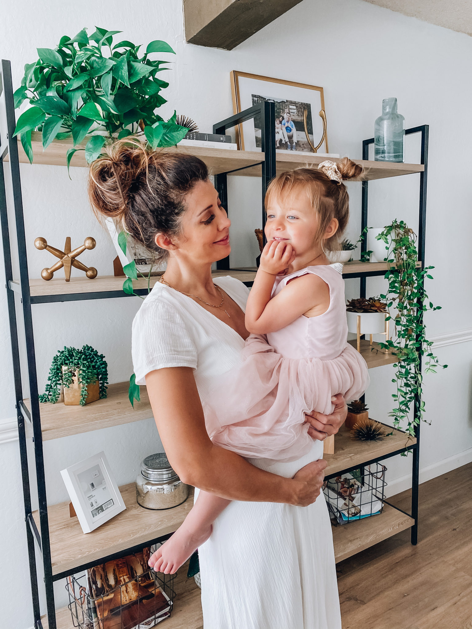 First Trimester Survival by popular San Francisco lifestyle blog, The Girl in the Yellow Dress: image of a woman wearing a white Nordstrom Blouson Dress WAYF and holding her young daughter as they standing in their home that's decorated with a World Market Natural Macrame Table Runner, Target 72" Loring 5 Shelf Ladder Bookcase, Amazon Nearly Natural 4762 44in. Philodendron Hanging Basket Silk Plant, Target Ceramic Planter White, and CB2 SIDERA CHAIR.