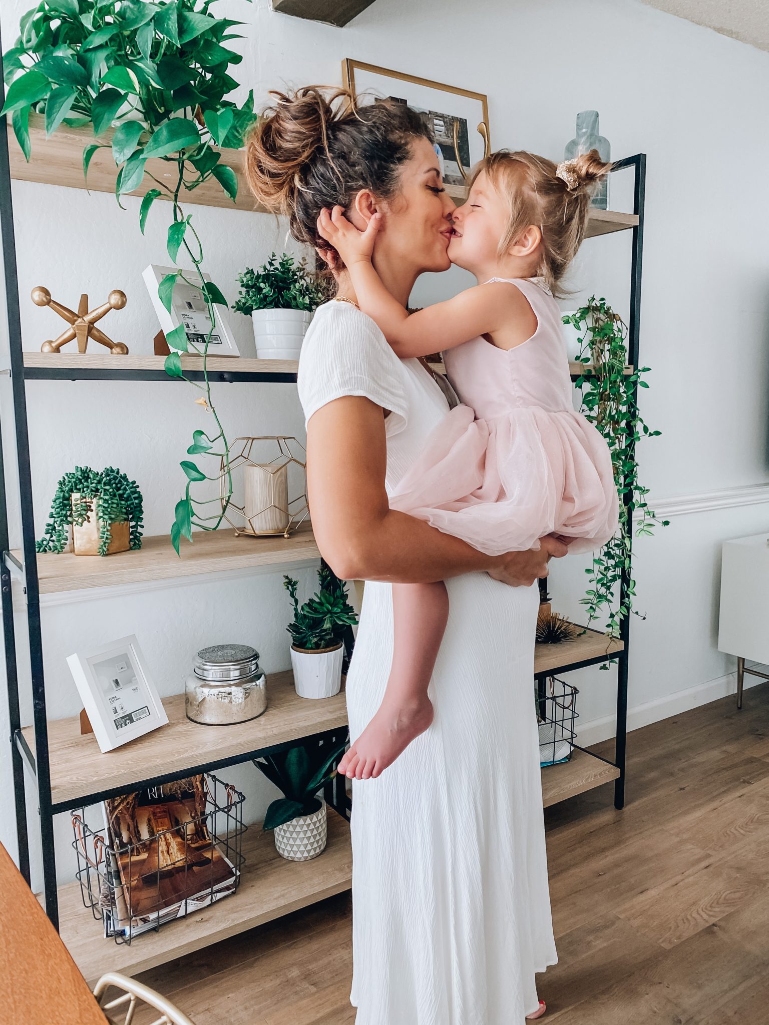 First Trimester Survival by popular San Francisco lifestyle blog, The Girl in the Yellow Dress: image of a woman wearing a white Nordstrom Blouson Dress WAYF and holding her young daughter as they standing in their home that's decorated with a World Market Natural Macrame Table Runner, Target 72" Loring 5 Shelf Ladder Bookcase, Amazon Nearly Natural 4762 44in. Philodendron Hanging Basket Silk Plant, Target Ceramic Planter White, and CB2 SIDERA CHAIR.