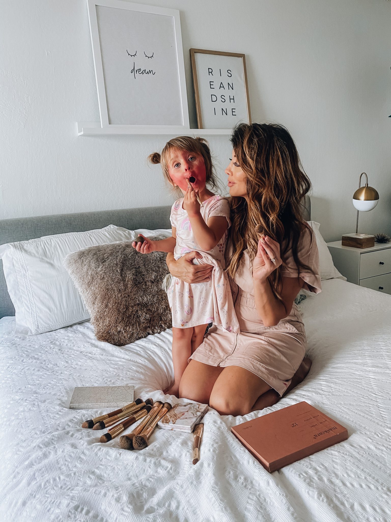 Maskcara Makeup by popular San Francisco beauty blog, The Girl in the Yellow Dress: image of a woman wearing a Target Short Sleeve V-Neck Zip-Up Elastic Waist Dress and sitting on her bed with her young daughter next to some Maskcara makeup pallets and Maskcara makeup brushes. 