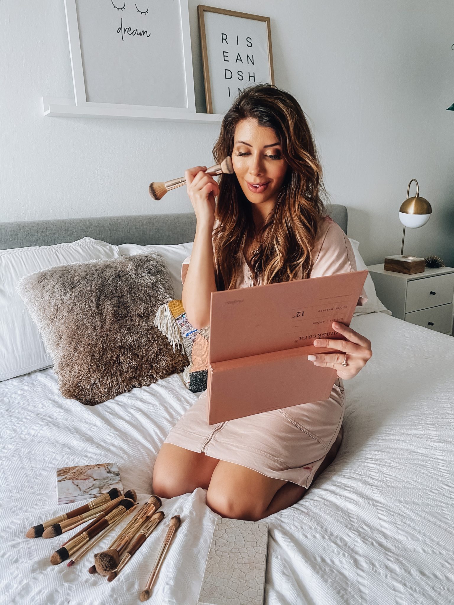 Maskcara Makeup by popular San Francisco beauty blog, The Girl in the Yellow Dress: image of a woman wearing a Target Short Sleeve V-Neck Zip-Up Elastic Waist Dress and sitting on her bed next to some Maskcara makeup palettes and Maskcara makeup brushes. 