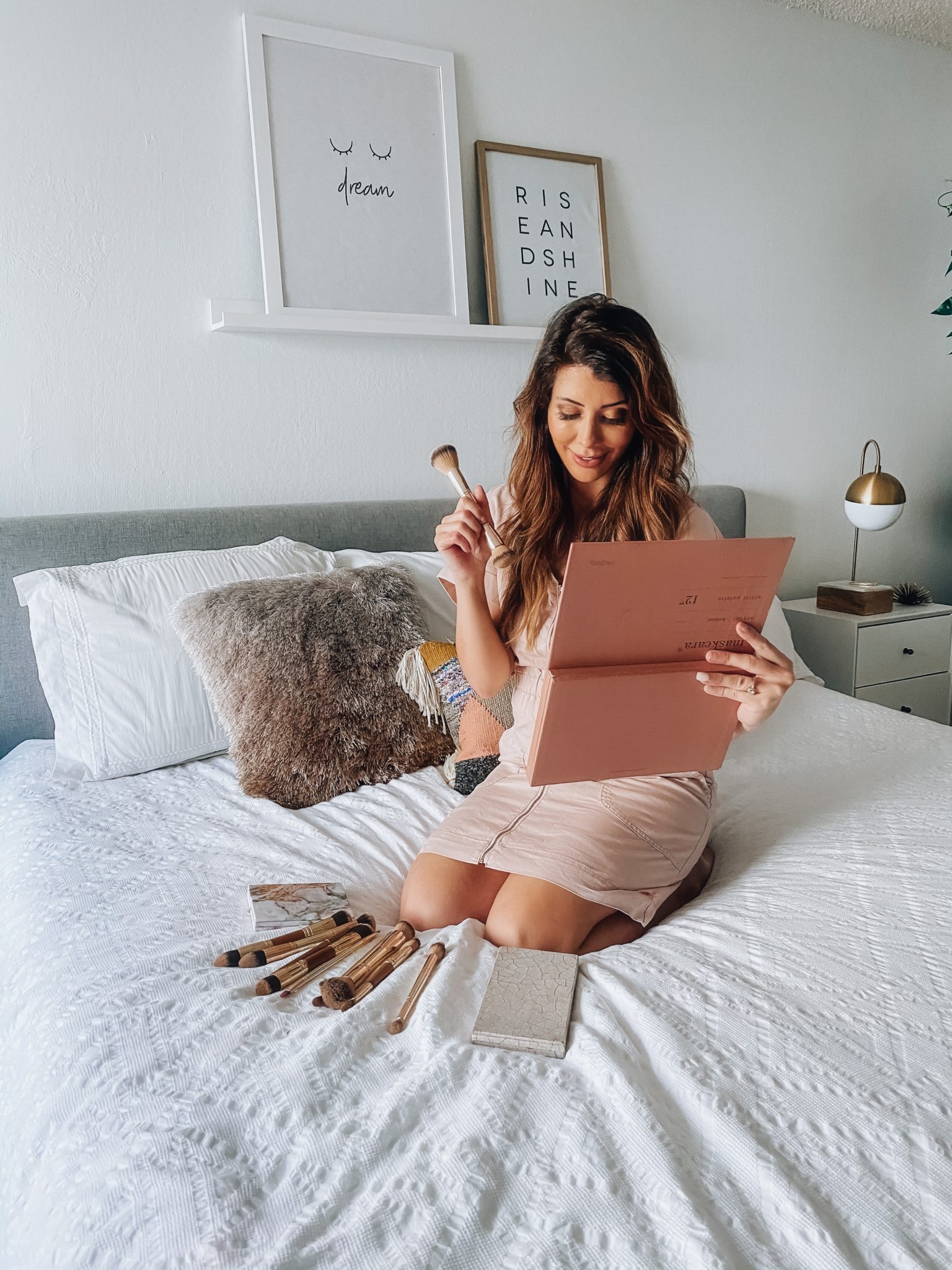 Maskcara Makeup by popular San Francisco beauty blog, The Girl in the Yellow Dress: image of a woman wearing a Target Short Sleeve V-Neck Zip-Up Elastic Waist Dress and sitting on her bed next to some Maskcara makeup palettes and Maskcara makeup brushes. 