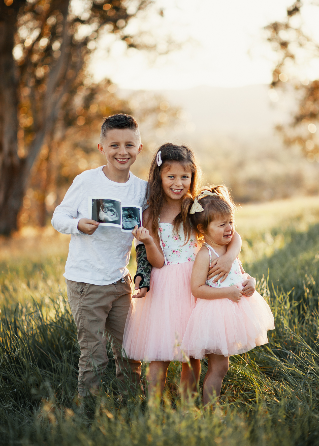 Family Pregnancy Announcement by popular San Francisco lifestyle blog, The Girl in the Yellow Dress: image of a brother and two sisters standing together in a field, holding some ultra sound pictures and wearing a Amazon Flofallzique Baby Girls Dress Toddler Tutu Infant Floral Sundress Tulle Wedding Dress, Freshly Picked White Patent Malibu, Old Navy Softest Crew-Neck Long-Sleeve Tee for Boys, and Old Navy Built-In Flex Twill Joggers for Boys.