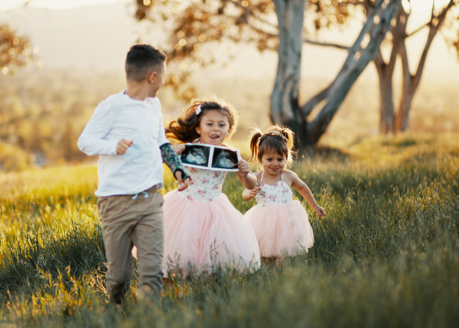 Family Pregnancy Announcement by popular San Francisco lifestyle blog, The Girl in the Yellow Dress: image of a boy and two girls running through a field, holding ultra sound photos and wearing Amazon Flofallzique Baby Girls Dress Toddler Tutu Infant Floral Sundress Tulle Wedding Dress, Freshly Picked White Patent Malibu, Old Navy Softest Crew-Neck Long-Sleeve Tee for Boys, and Old Navy Built-In Flex Twill Joggers for Boys.