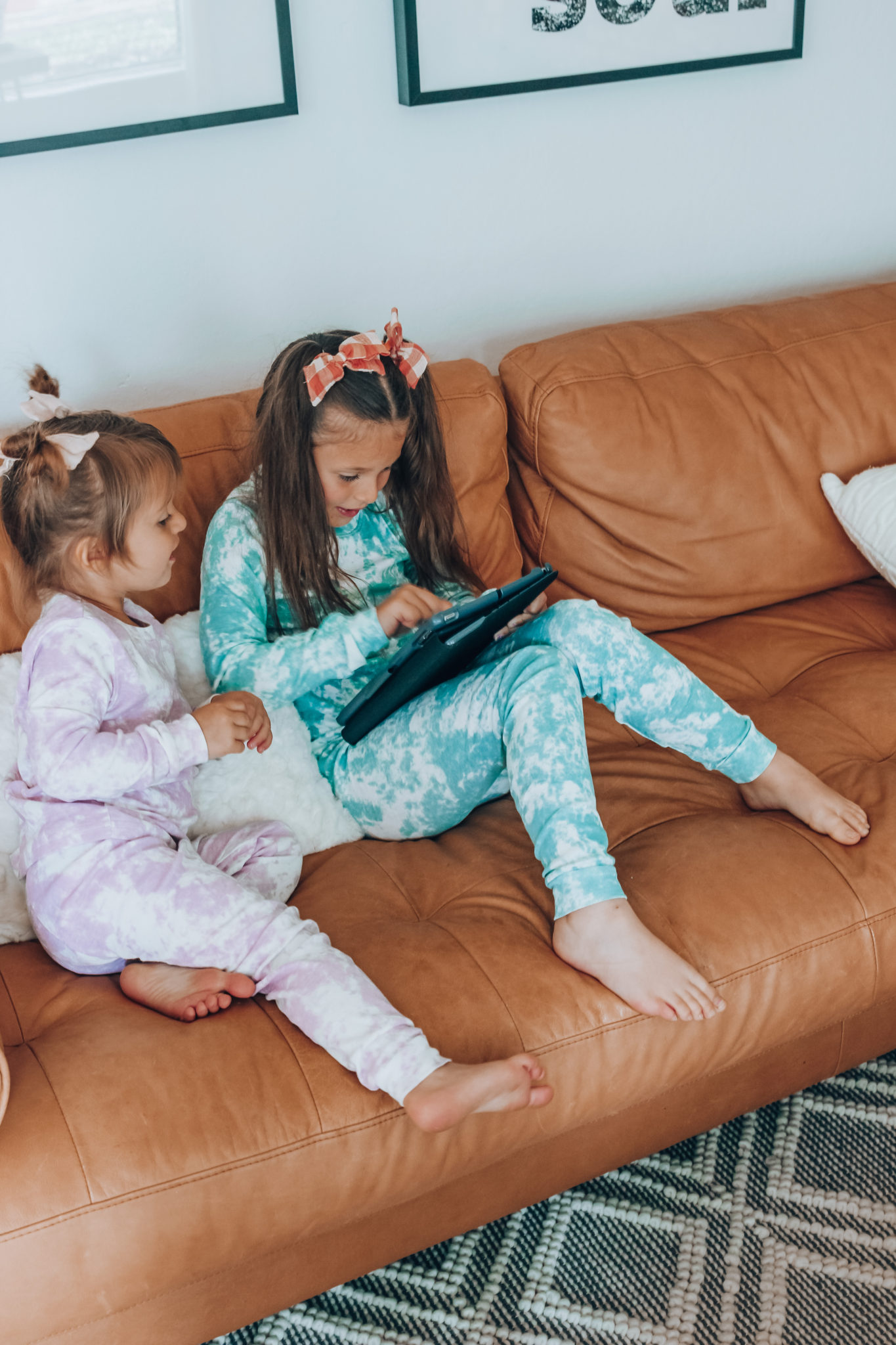 Messenger Kids Review by popular San Francisco lifestyle blog,The Girl in the Yellow Dress: image of two young girls wearing tie dye pajamas and sitting together on a leather couch while they look at a tablet. 